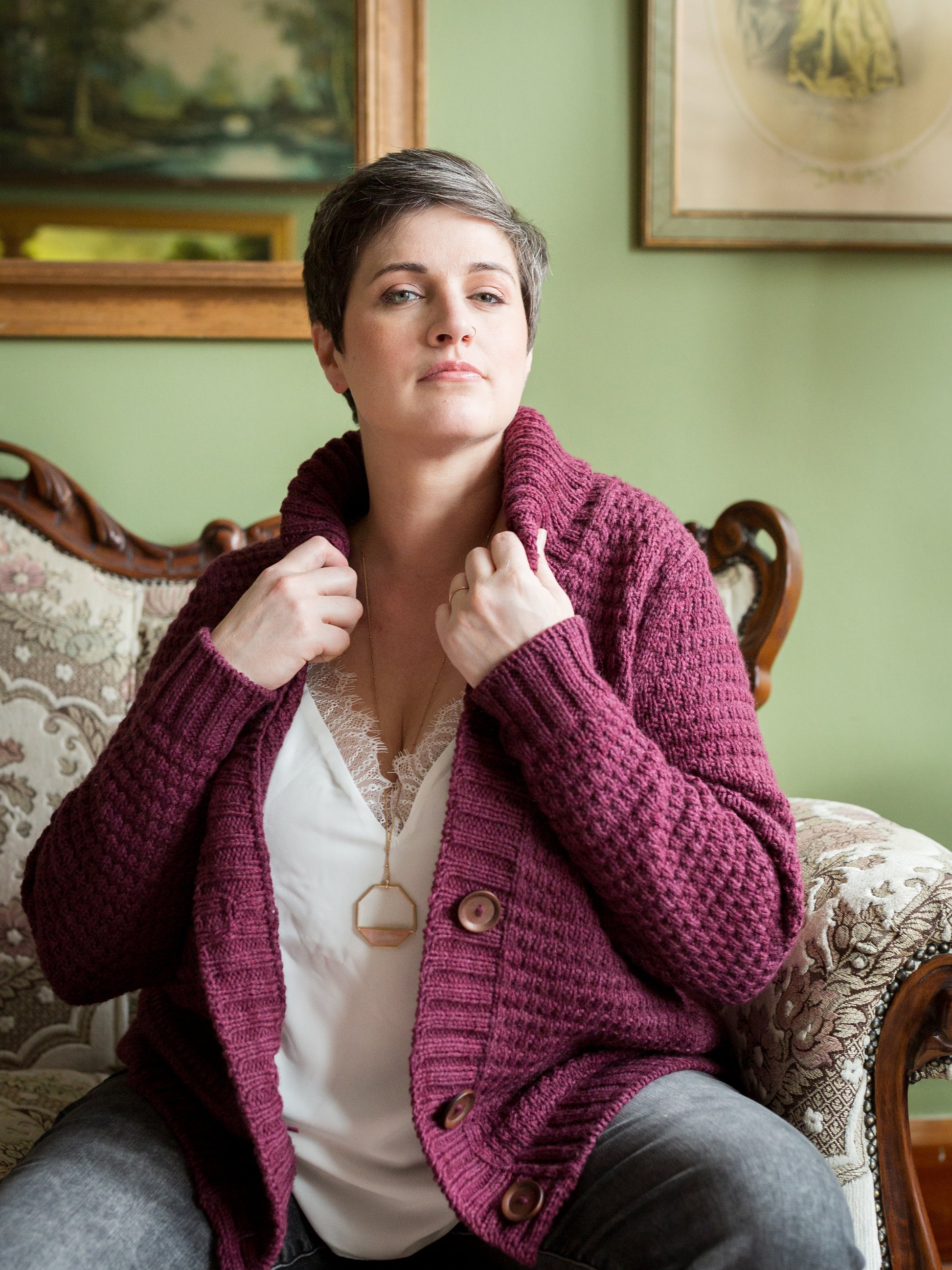 Jen sits on an antique couch, wearing a hand knit cardigan with a thick button band and a cowl neck. She looks into the camera.