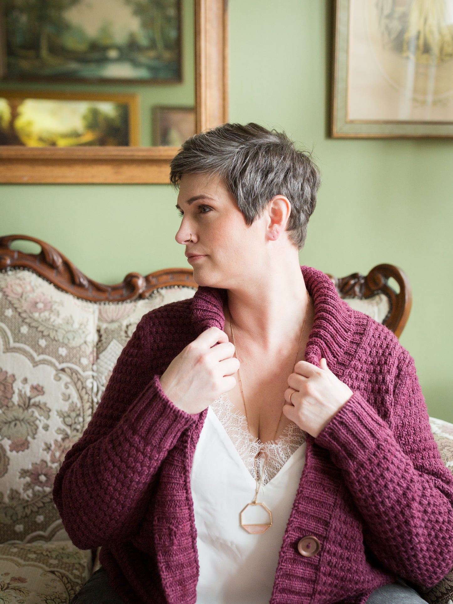 Jen sits in front of the camera, looking to the side. Over a white camisole, she wears a hand knit burgundy cardigan. She adjusts the thick cowl neck, showing off the ribbing detail.