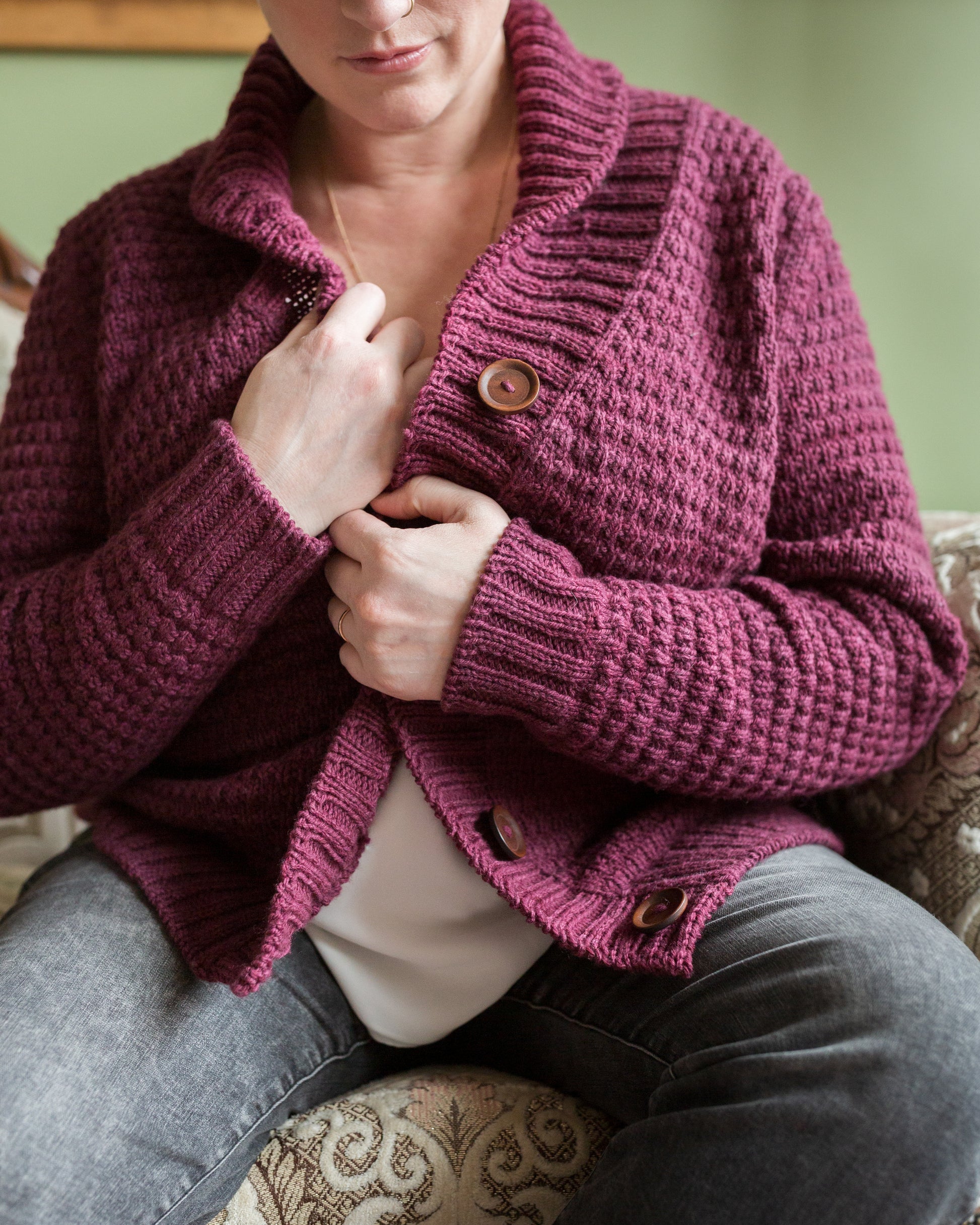 Seen close up, Jen pulls the sides of her hand knit cardigan together. The cardigan has a cowl neck and a large ribbed button band, alongside a subtle all-over stitch detail.