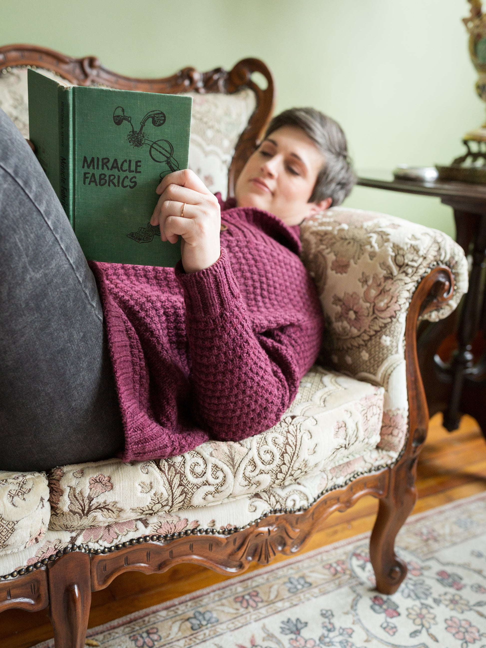 Jen reclines on a couch, reading a copy of Miracle Fabrics. She wears a pair of black jeans with a burgundy moss stitch cardigan, knit from her Postscript cardigan pattern.