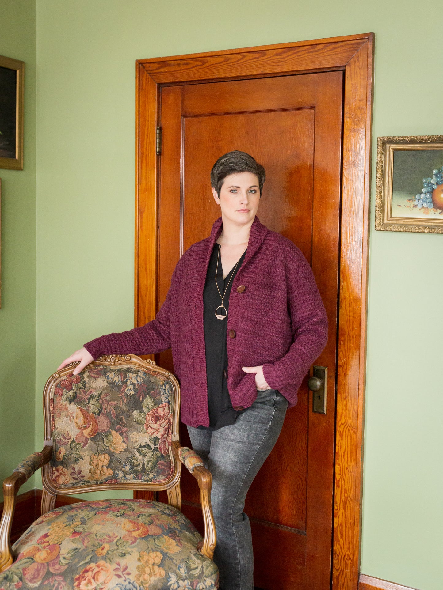 Jen stands, one hand on an antique chair and the other in the pocket of her grey jeans, looking at the camera. She wears a burgundy cardigan, knit with a cowl neck and a large button band.