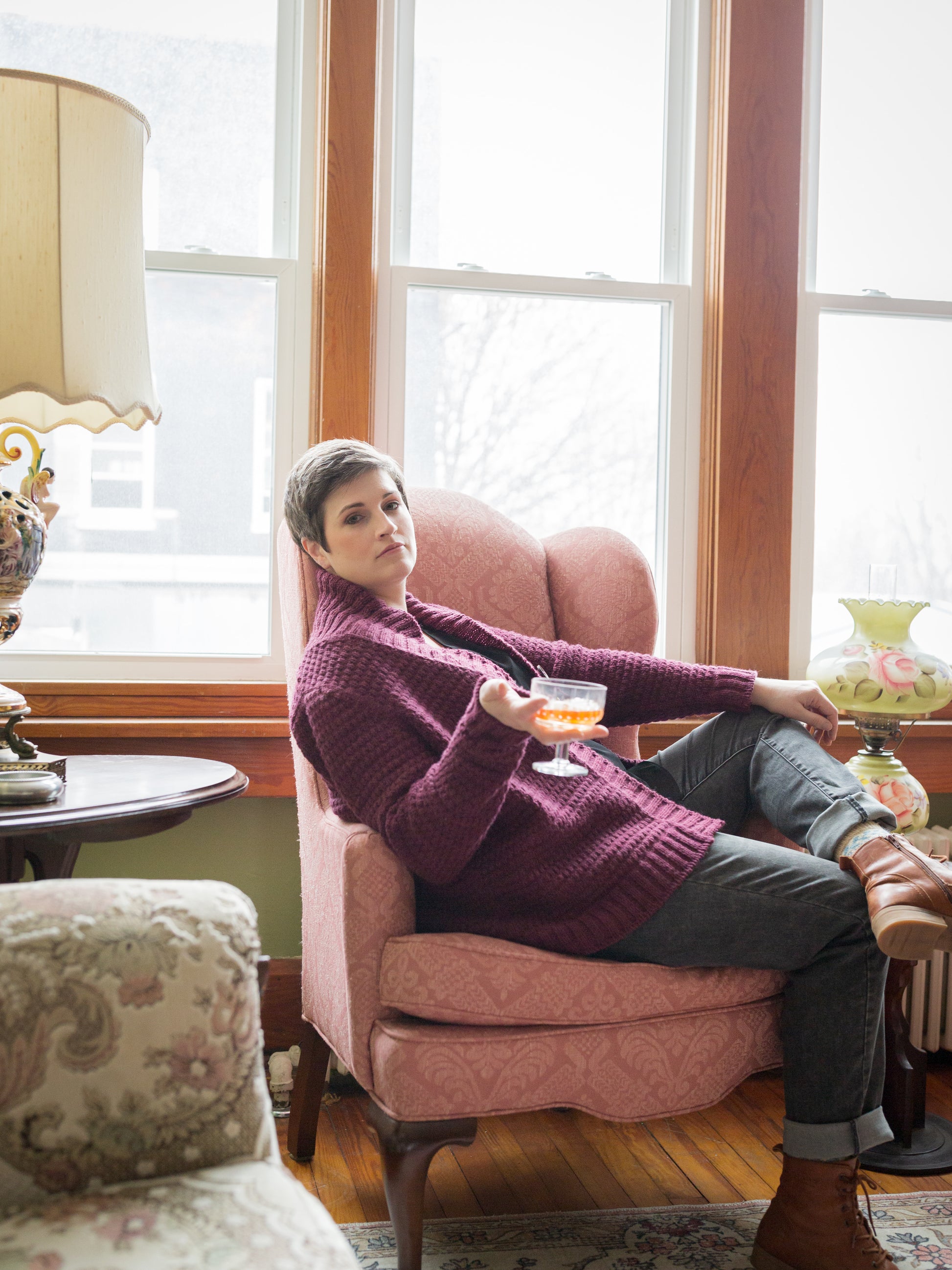 Jen reclines in an antique chair, holding a coupe glass in one hand. She wears brown boots and grey jeans with a burgundy cardigan that's knit with a cowl neck, ribbed button band, and all-over stitch pattern.