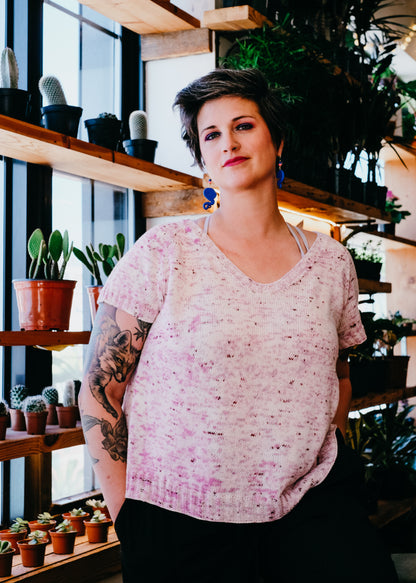 Jen stands, looking at the camera. She wears a hand knit tee, made with light pink speckled yarn, with black pants. Houseplants can be seen in the background.