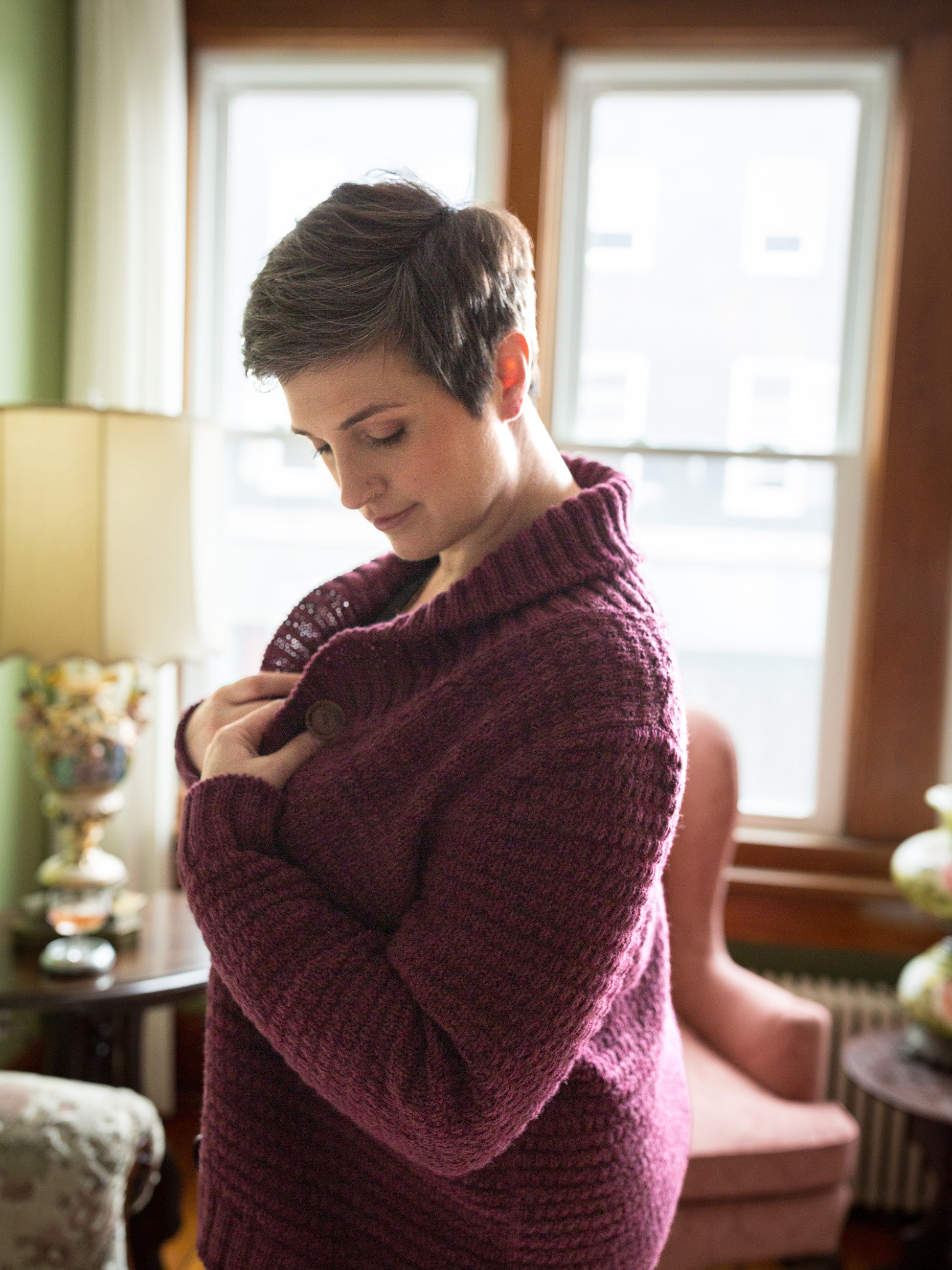 Seen from the side, Jen wears a cardigan, knit with a cowl neck and a subtle allover stitch. A window and an antique lamp can be seen in the background.