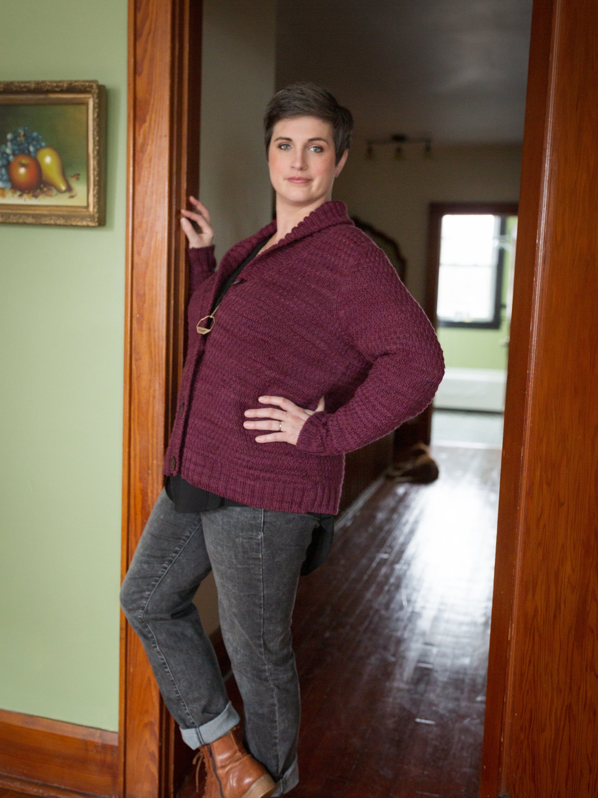 Jen stands in the doorway, at an angle to the camera. She has one hand on her hip, showing off the masculine structure of the hand knit, burgundy cardigan that she wears with brown boots and grey jeans.