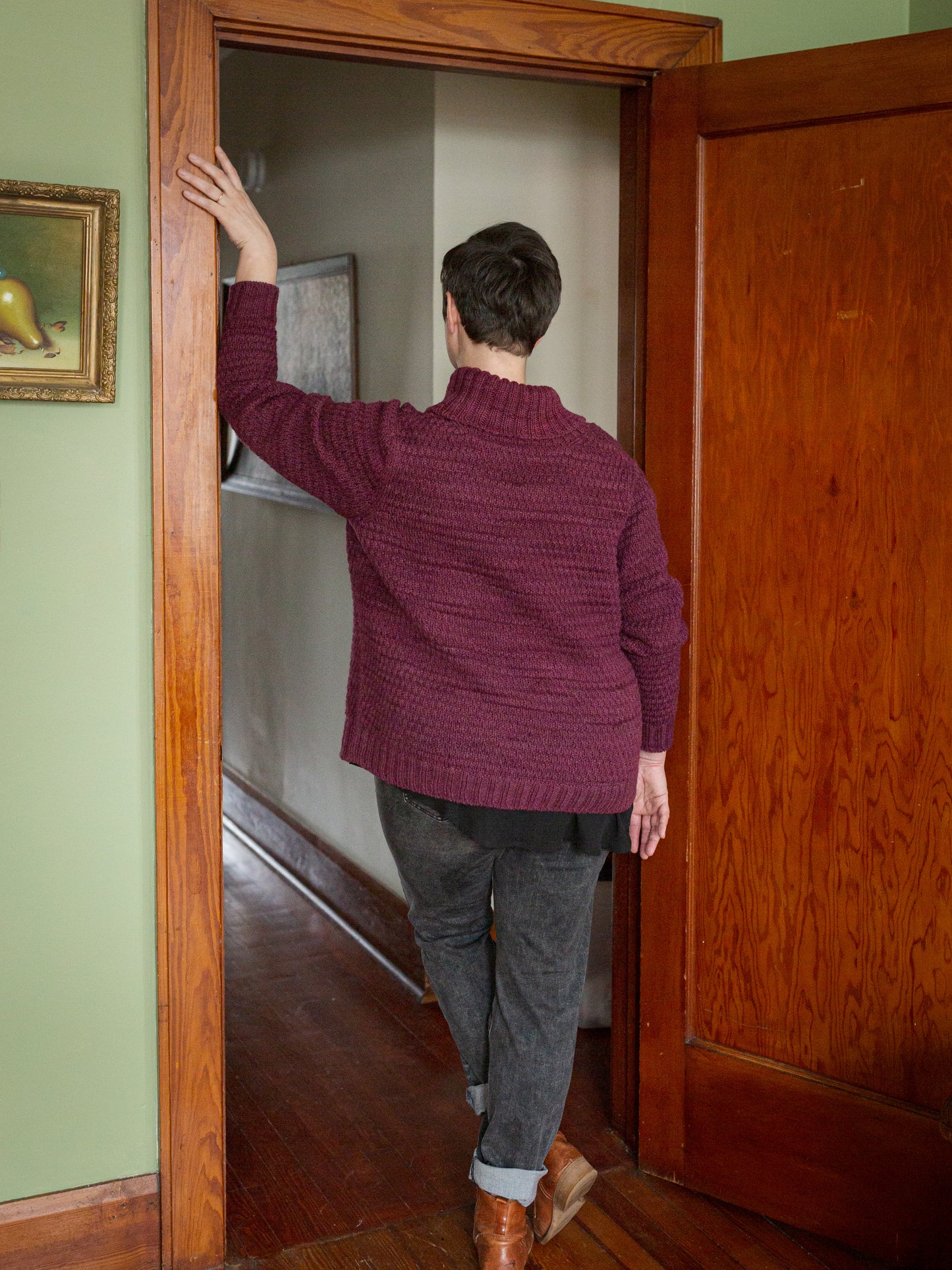 Seen from behind, Jen stands in a doorway, one arm raised to show off the structured, hand knit cardigan she wears with grey jeans and brown boots.