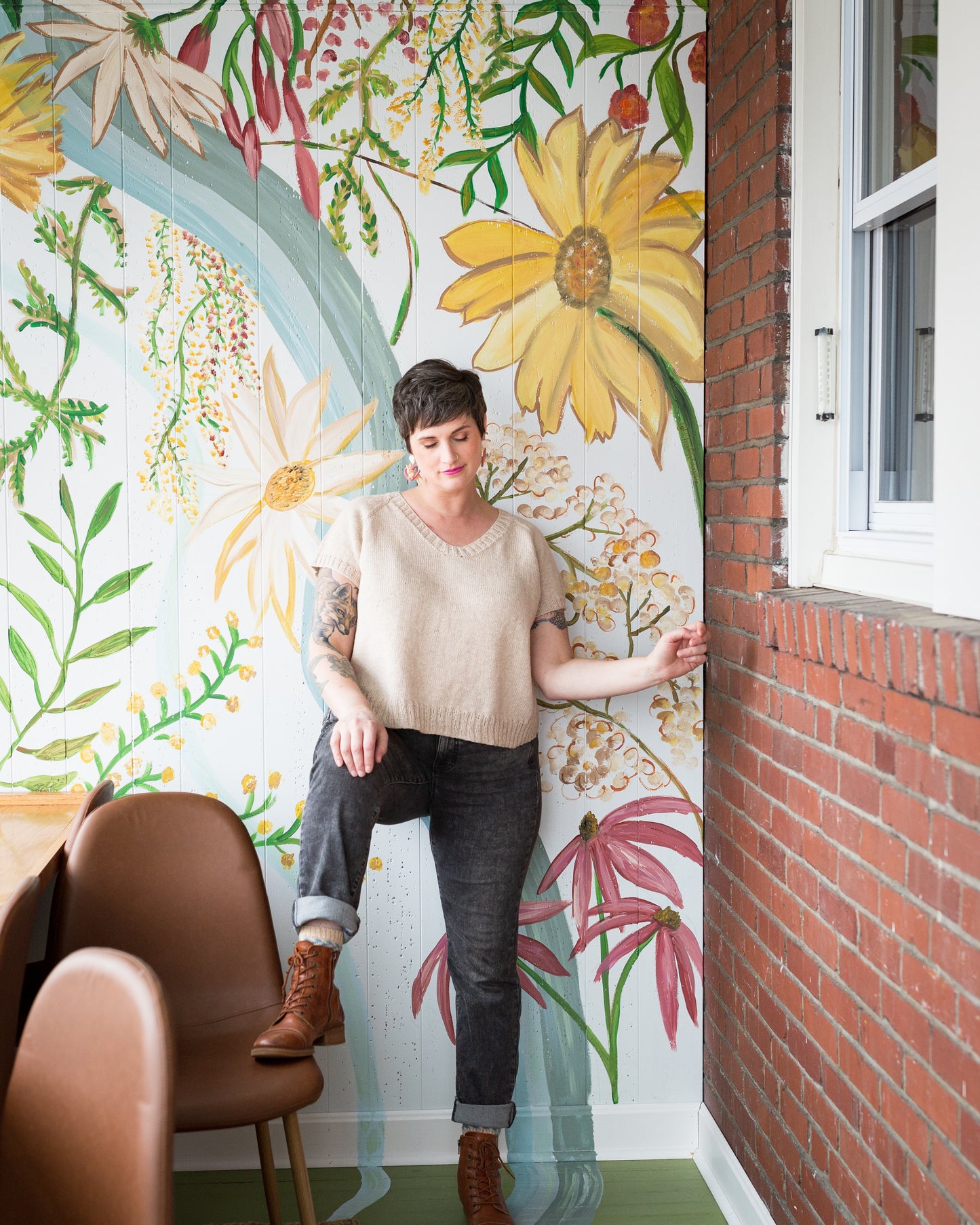 Jen stands, one foot up on a chair, looking down. She wears a light beige tee, knit with a V-neck, and gray jeans with brown boots. A floral wall mural can be seen in the background.
