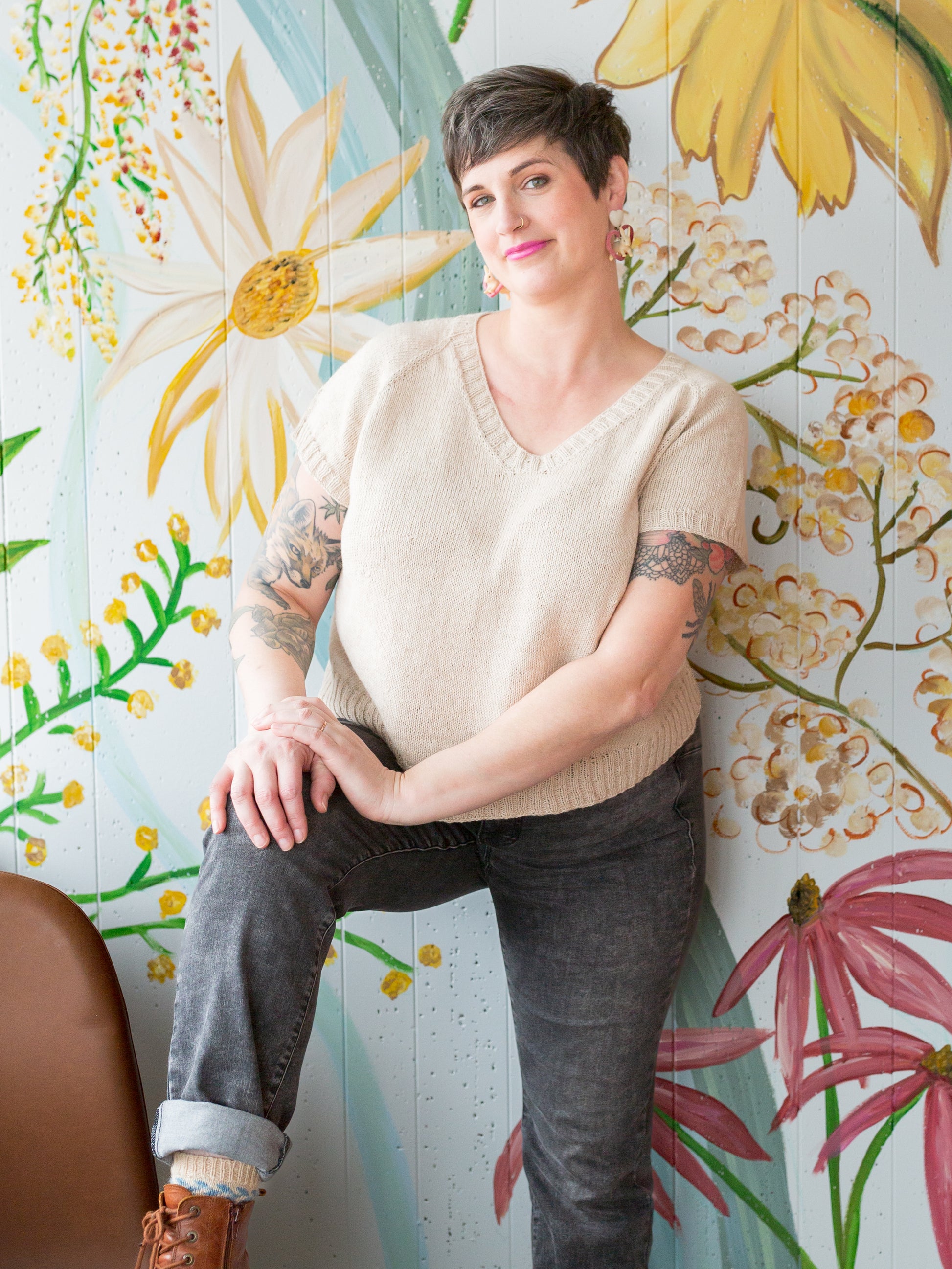 Jen stands, smiling at the camera, wearing a tee knit with light beige yarn with grey jeans and brown boots. A floral wall mural can be seen in the background.