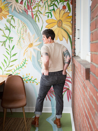 Seen from behind, Jen wears a light beige, hand knit tee with gray jeans and brown boots. A floral wall mural and brick can be seen in the background.