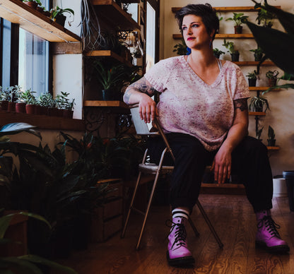 Jen sits on a chair, looking off camera. She wears pink boots, black pants, and a light pink tee. The tee is knit with a gentle V-neck.