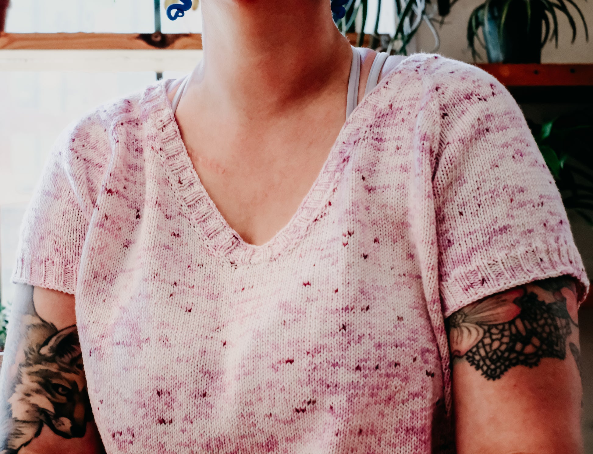 Seen close up, Jen wears a light pink, speckled tee, knit with a gentle V-neck. Houseplants can be seen in the background.