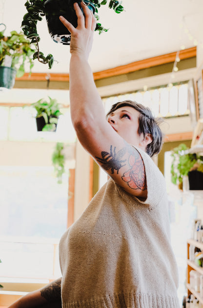 Jen holds up a hanging houseplant, wearing a light beige, hand knit tee.