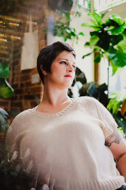 Jen leans back, surrounded by houseplants, wearing a light beige, hand knit tee. She looks off camera.