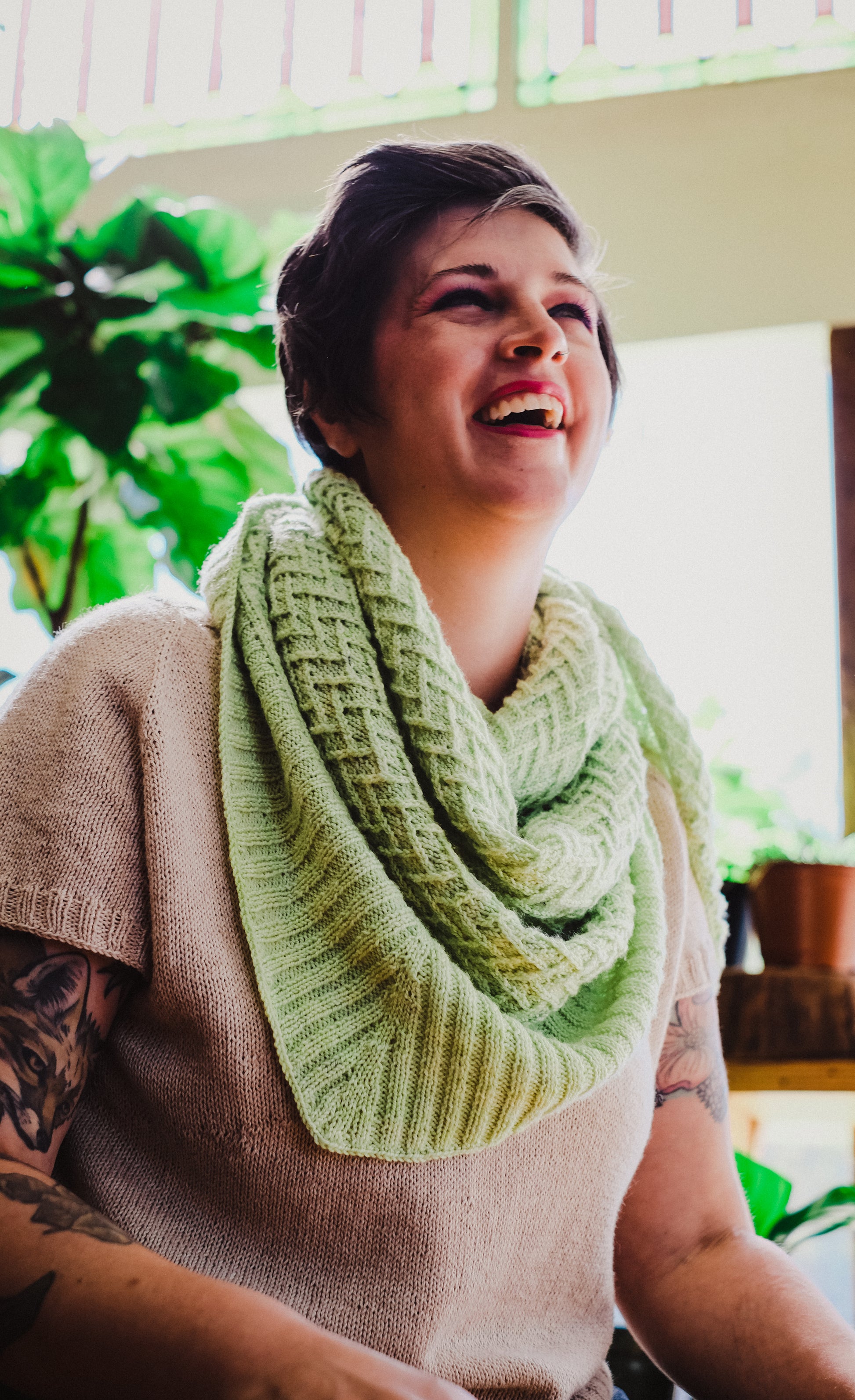 Jen looks off camera, laughing. She wears a lime green knit shawl with chevron details over a hand knit, light pink tee.