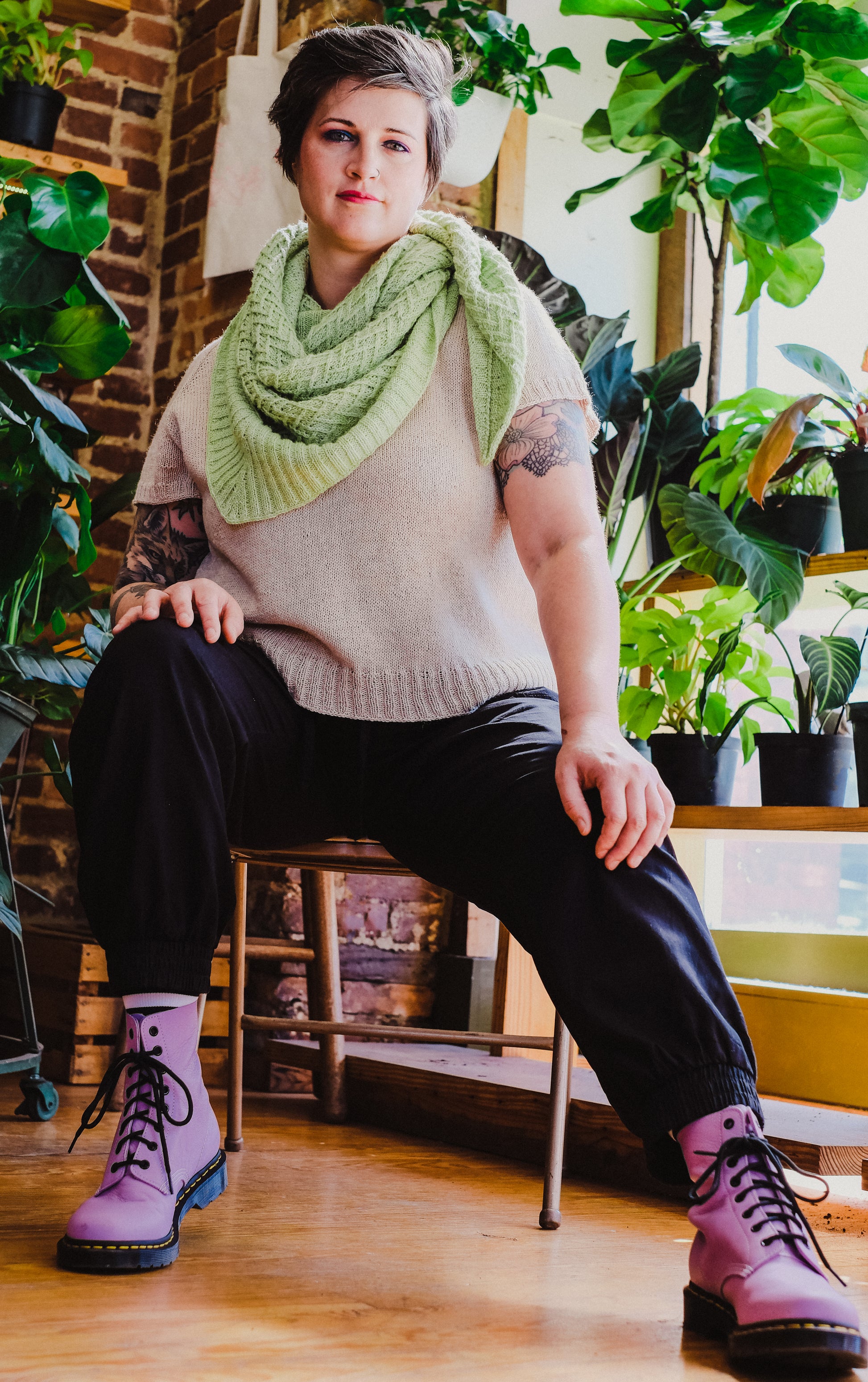 Jen sits on a chair, looking down at the camera. She wears a neon green scarf - the Sepal shawl - with a light beige, hand knit tee and black pants. Houseplants can be seen in the background.
