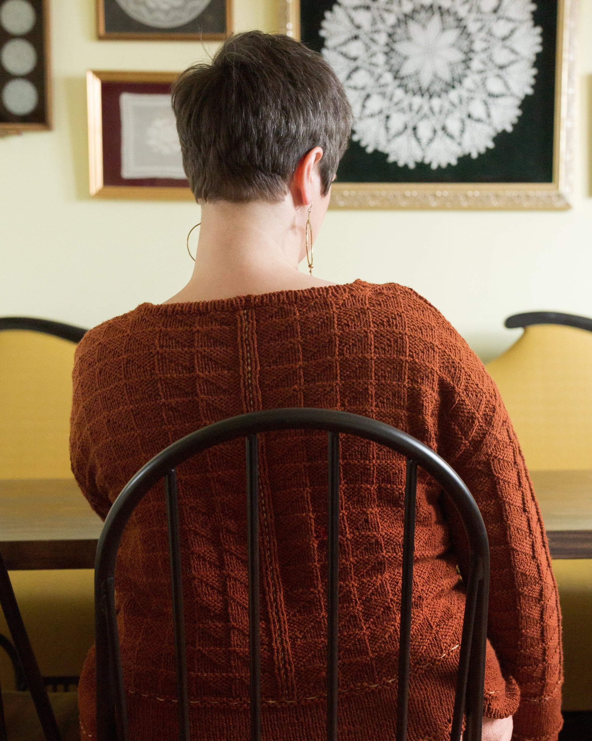 Seen from behind, Jen wears a knit pullover, made from brick orange yarn. The pullover features a allover quilt stitch design and a contrasting running stitch line down the back.