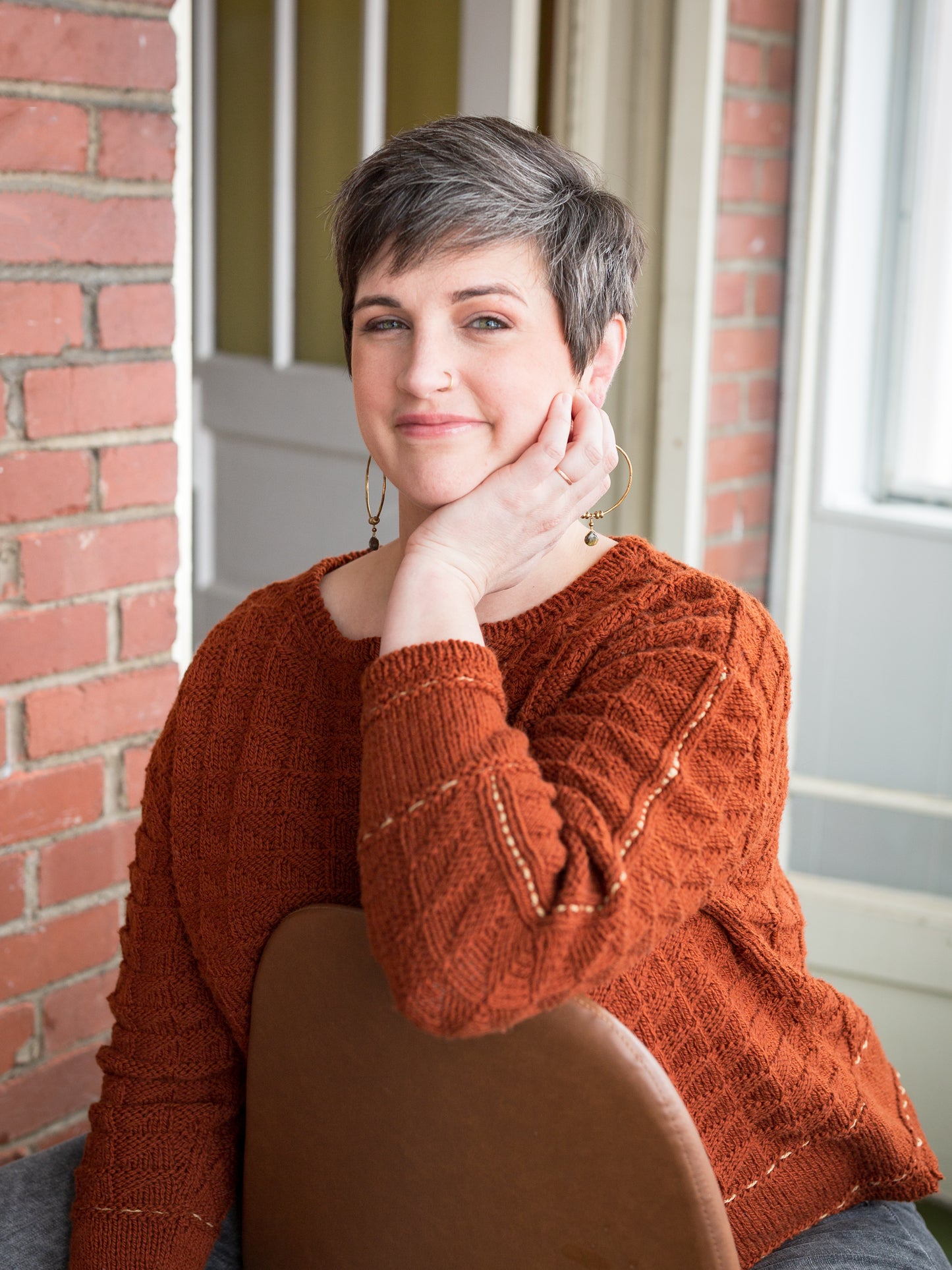 Jen sits in front of a brick wall, smiling at the camera. She wears a knit pullover with a quilt stitch texture that has a running stitch highlight in contrasting thread on the hems and arms. 