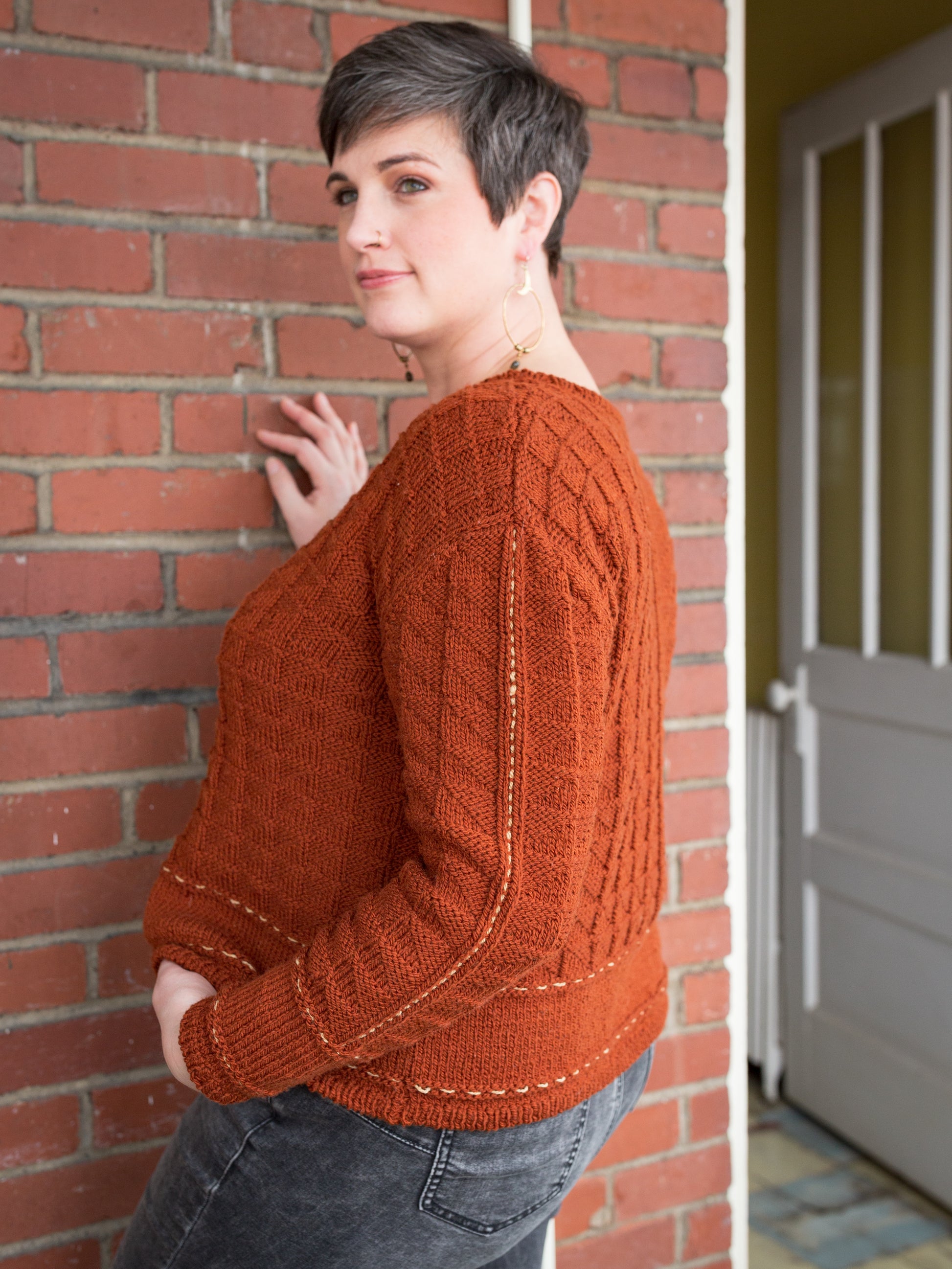 Seen from behind at a 3/4 angle, Jen wears a brick orange sweater, knit with an allover texture and featuring a contrasting running stitch on the arm and hem, with a pair of grey jeans. 