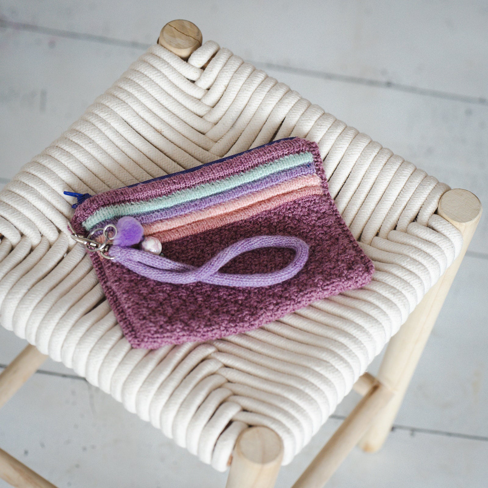 A zippered notions pouch has been hand knit from pink, green, orange, and purple fabric. It has a handle attached with a lobster claw clasp.