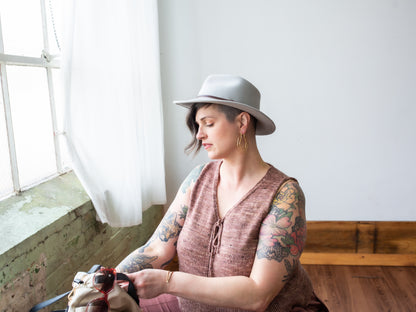 Jen sits, holding a white and black bag. She wears a dusty pink tank top with a bow tied in the front. The top was knit using the Ruffle Addendum pattern.