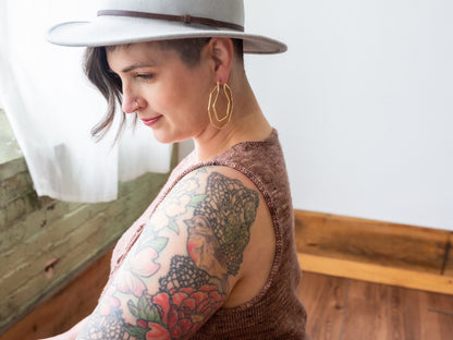 Seen from behind at an angle, Jen wears a dusty pink knit top with gold earrings and a white hat.