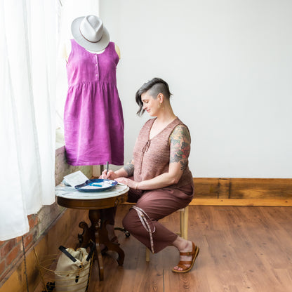 Jen sits at a table, wearing a pair of pink pants that match her hand knit pink tank top. Her dress form can be seen in the background.