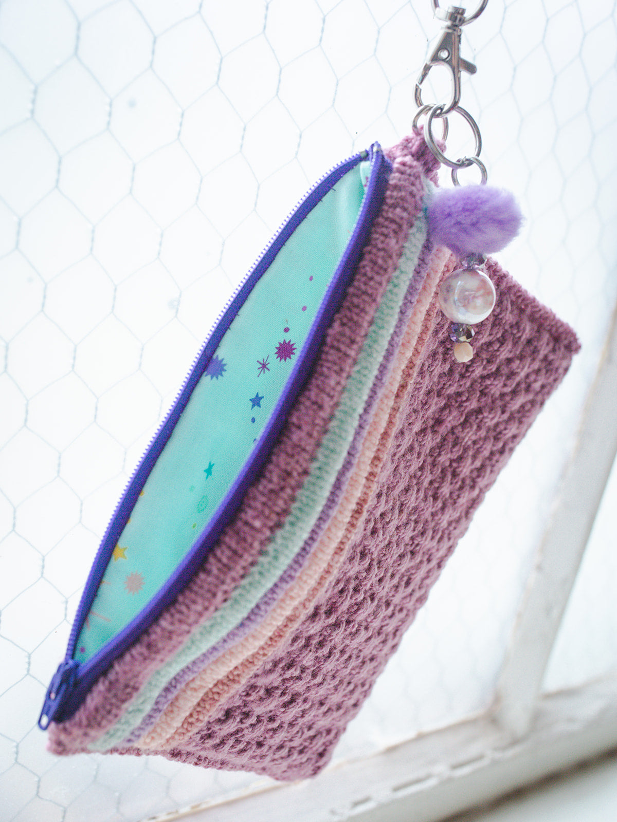 A pouch, lined with blue fabric, is knit out of pink fabrics. It has a lobster claw clasp and a keychain charm hanging off it.