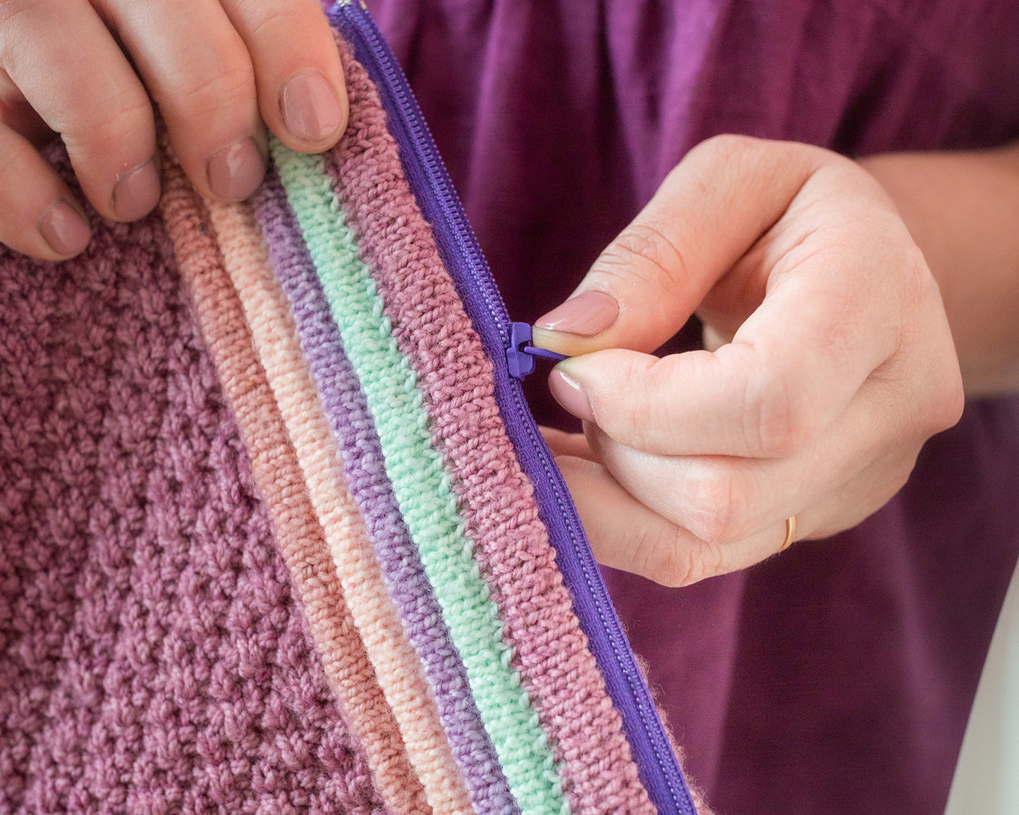 Jen's hands hold a Ziplet pouch, knit with dark pink, green, purple, and orange yarn. It has a purple zipper that Jen is unzipping.