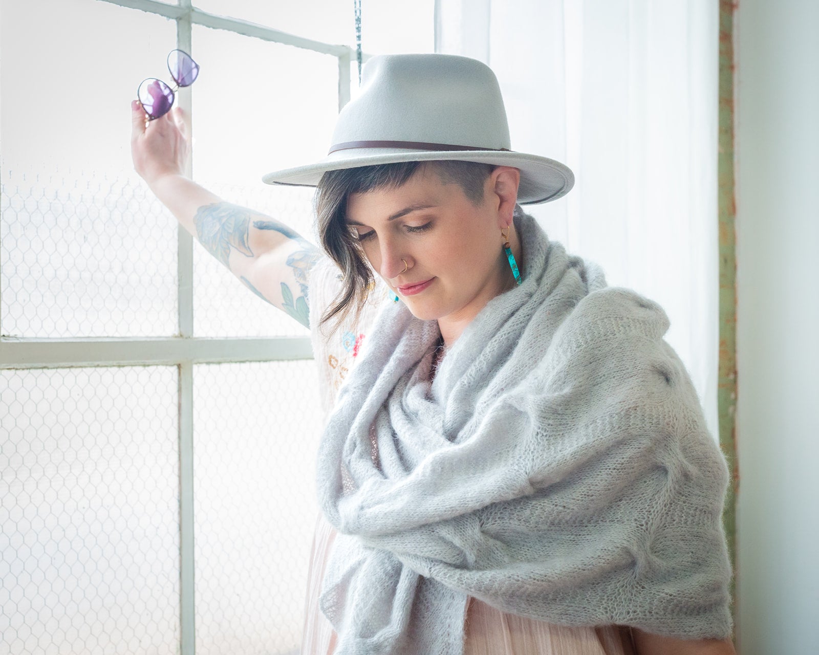 Jen, one hand against a window, wears a light blue, cable knit shawl with a white hat and a pink dress.