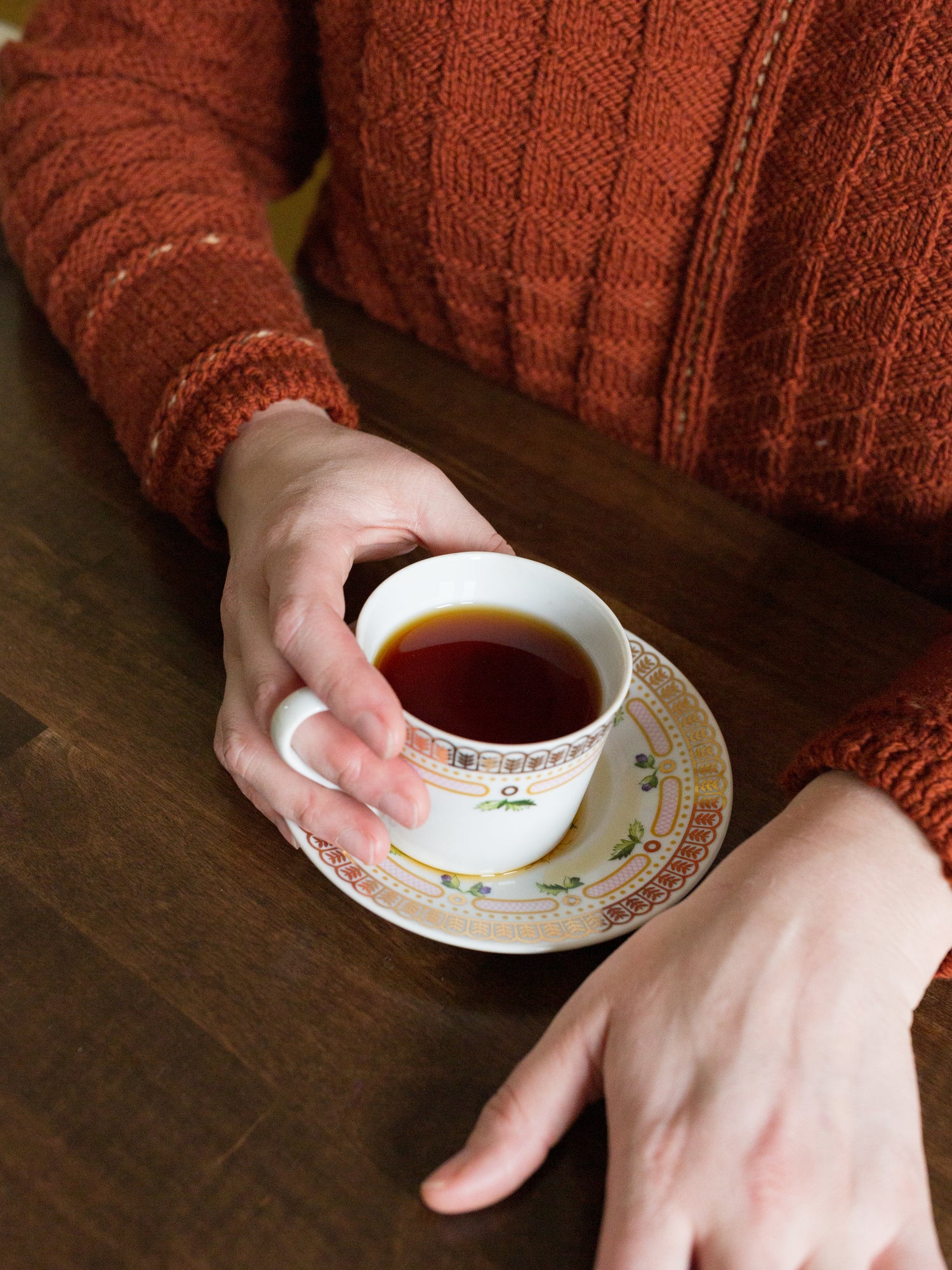Jen's hand holds a teacup and saucer. She wears her Maker's Tunic, a brick red handknit sweater.