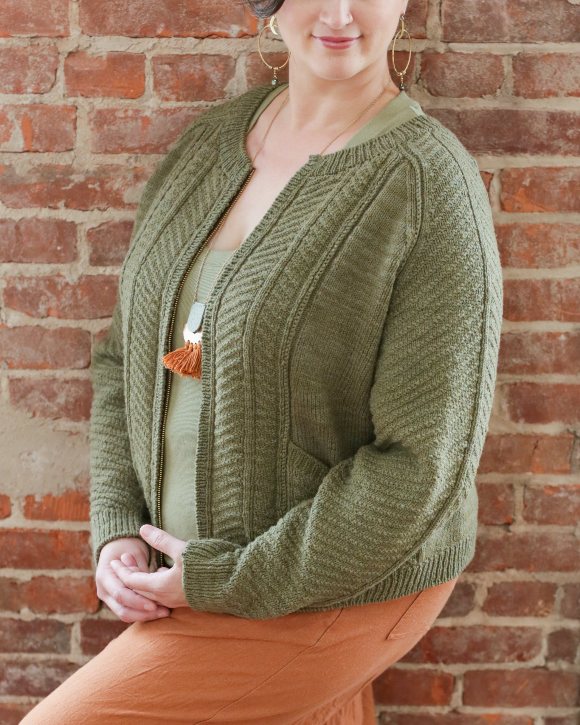 Seen from the neck down, Jen wears a green cardigan, knit with a large chevron pattern and a thin ribbed edging. The cardigan has two slanted front pockets and a zipper.