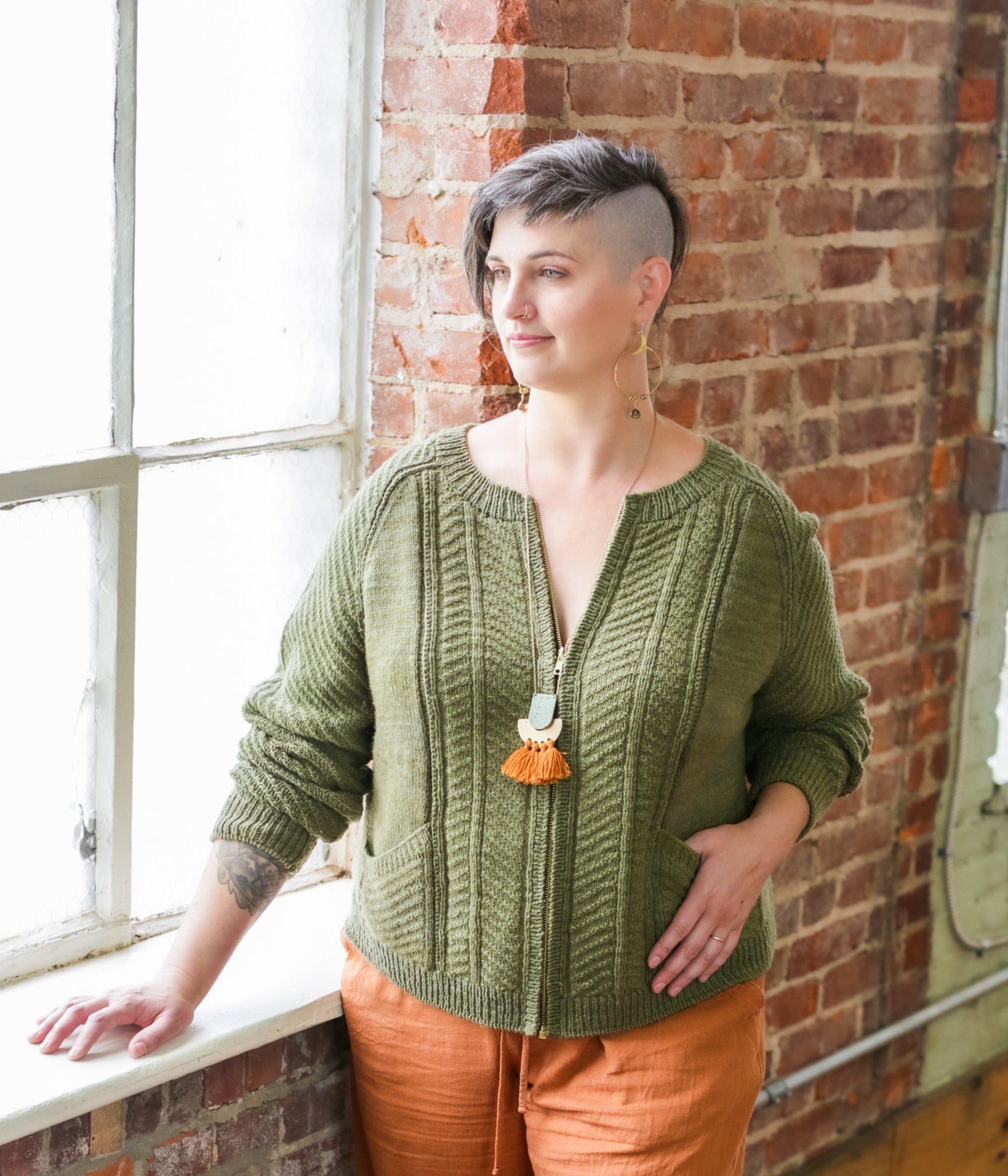 Jen stands next to a window, wearing orange pants with a partially zipped cardigan. The cardigan, knit in green yarn, has two slanted front pockets and a large chevron pattern down the front and arms.