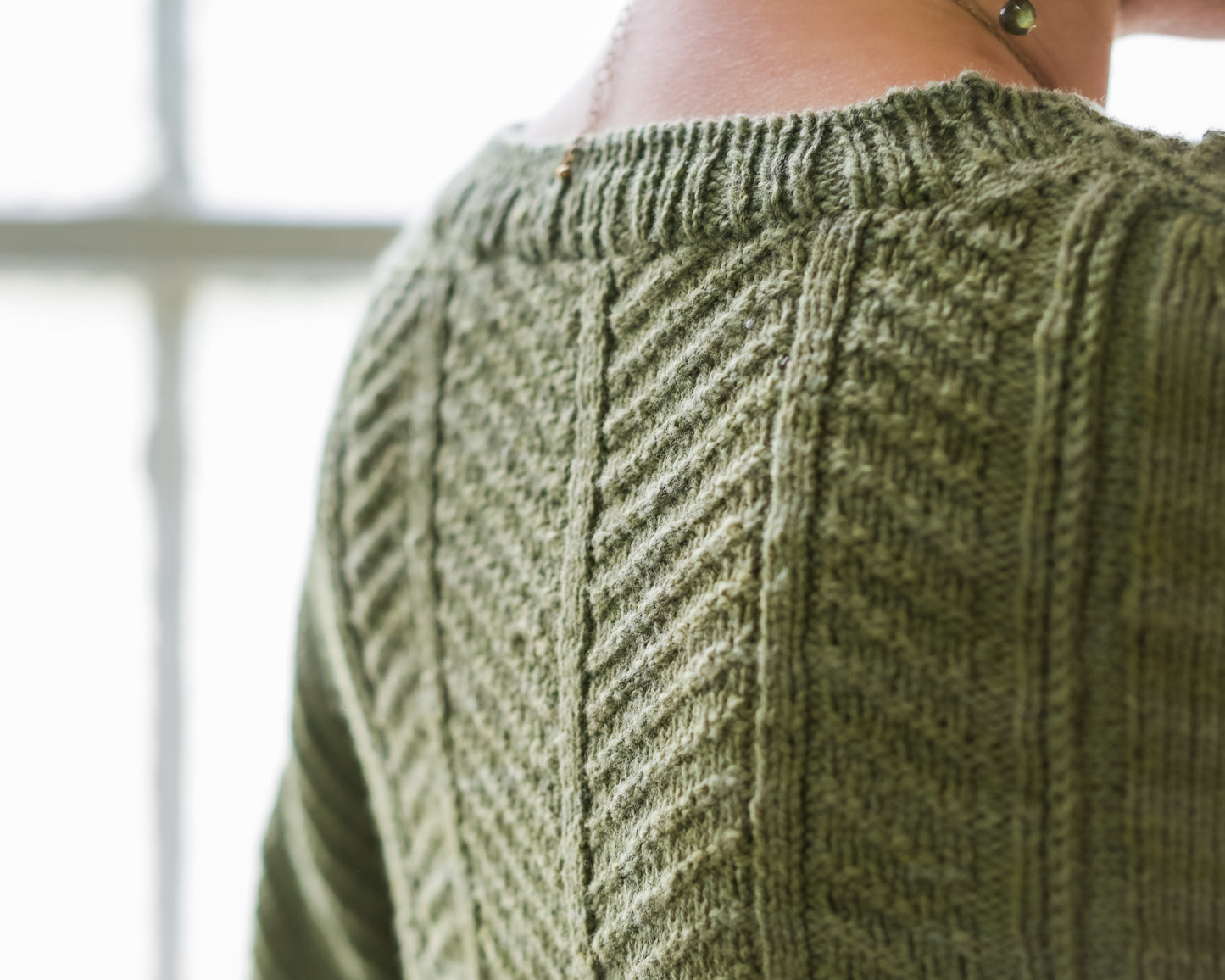 Seen up close front behind, Jen wears a hand knit cardigan. Made from green yarn, it features a thin ribbed edge and a central chevron design down the back.