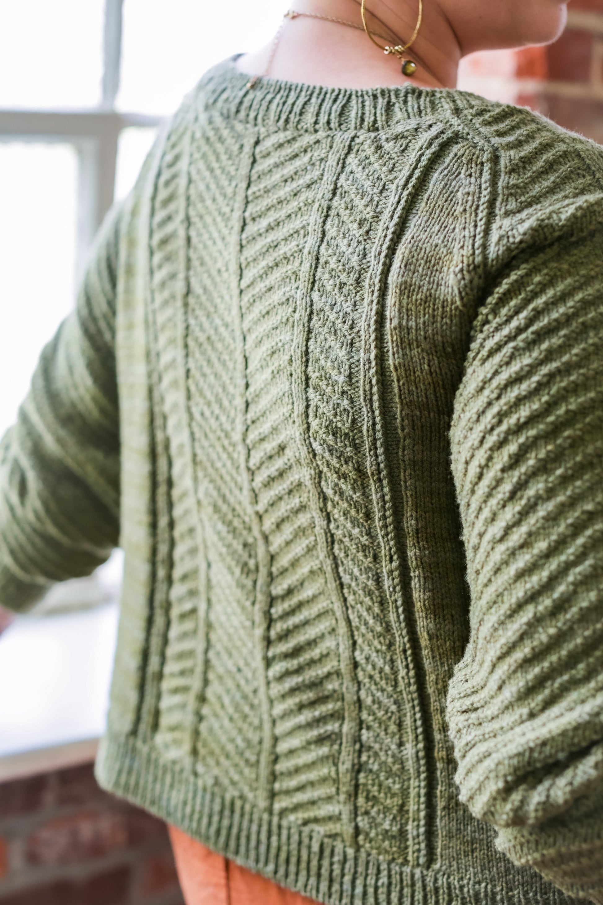 Seen from behind, Jen wears a green cardigan with orange pants. The cardigan has a thin ribbed edge and a central chevron pattern down the back, as well as chevron stitch down the arms. A window can be seen in the background.
