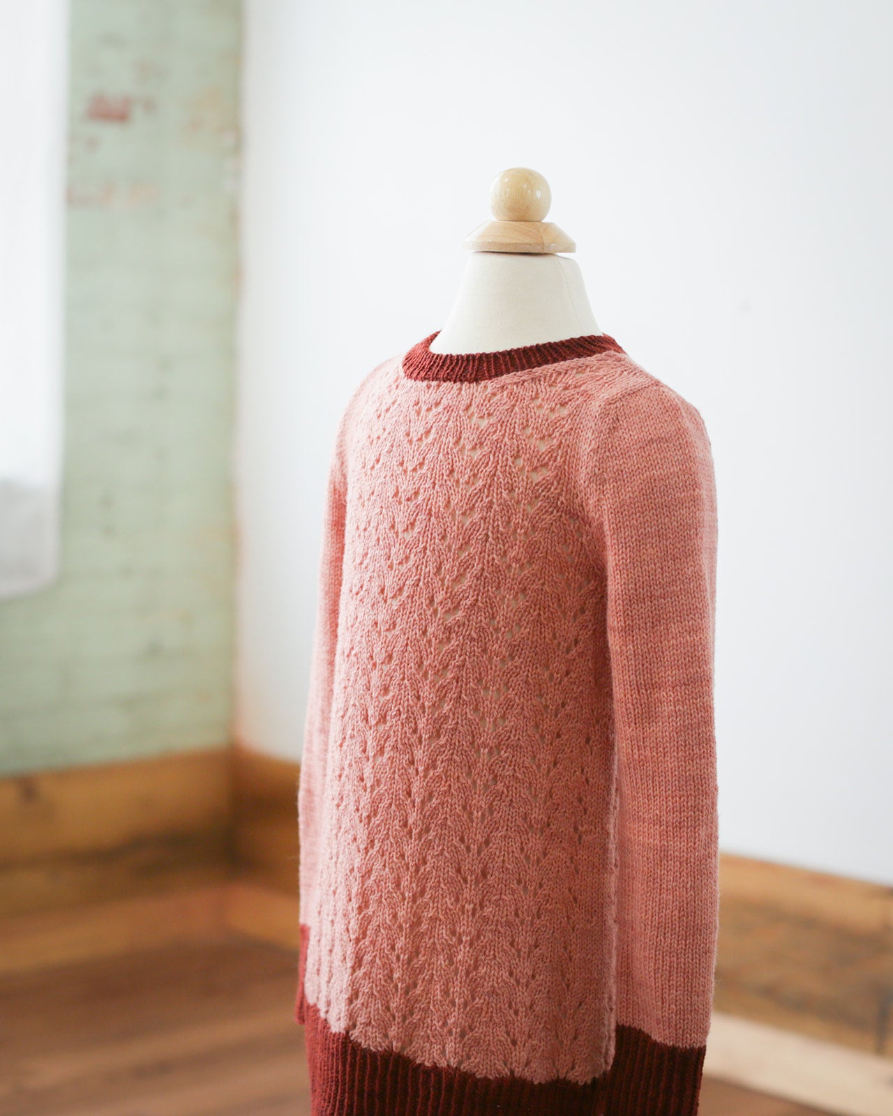 A dress length version of the Kid's Classic sweater - knit in light pink yarn from the lace body and dark pink yarn for the cuffs, hem, and collar - hangs on a dress form.
