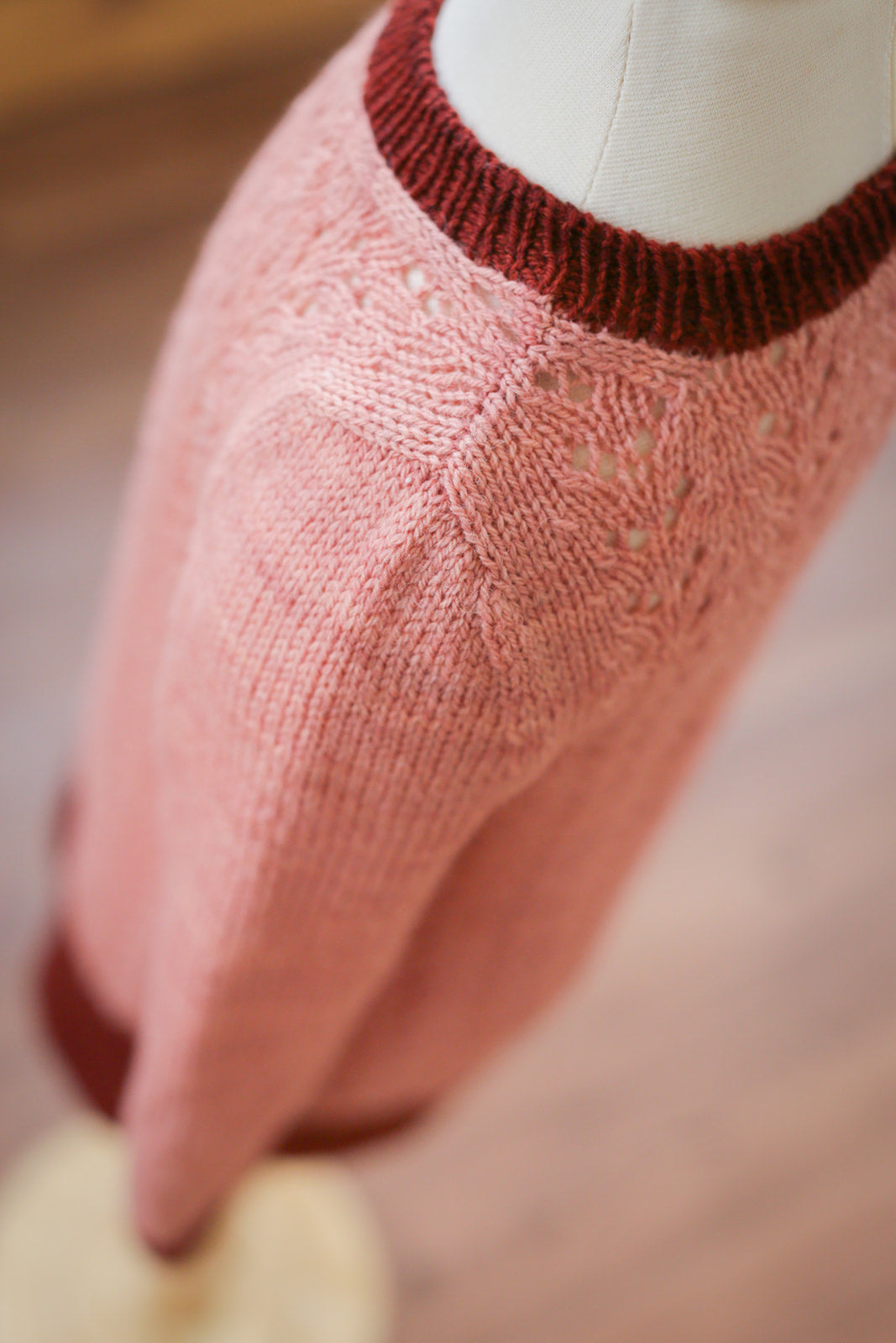 Seen from above, a dress length version of the Kid's Classic sweater - knit in light pink yarn from the lace body and dark pink yarn for the cuffs, hem, and collar - hangs on a dress form. The camera focuses on where the lace detail on the front and back meet at the shoulder seam.