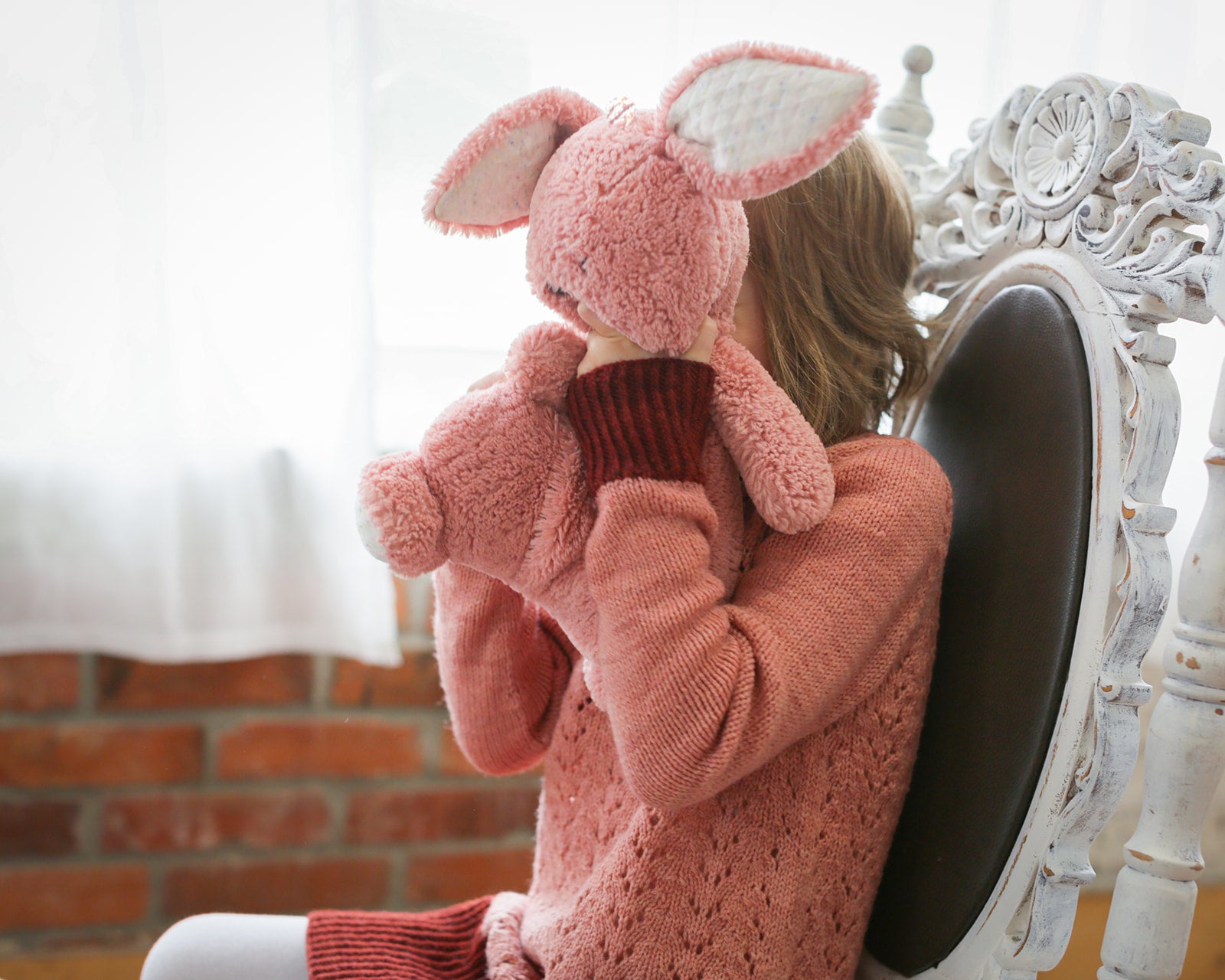 A young girl sits on a white antique chair, holding a pink stuffed animal up in front of her face. She wears a light pink knit dress, featuring dark pink cuffs, hem, and collar.