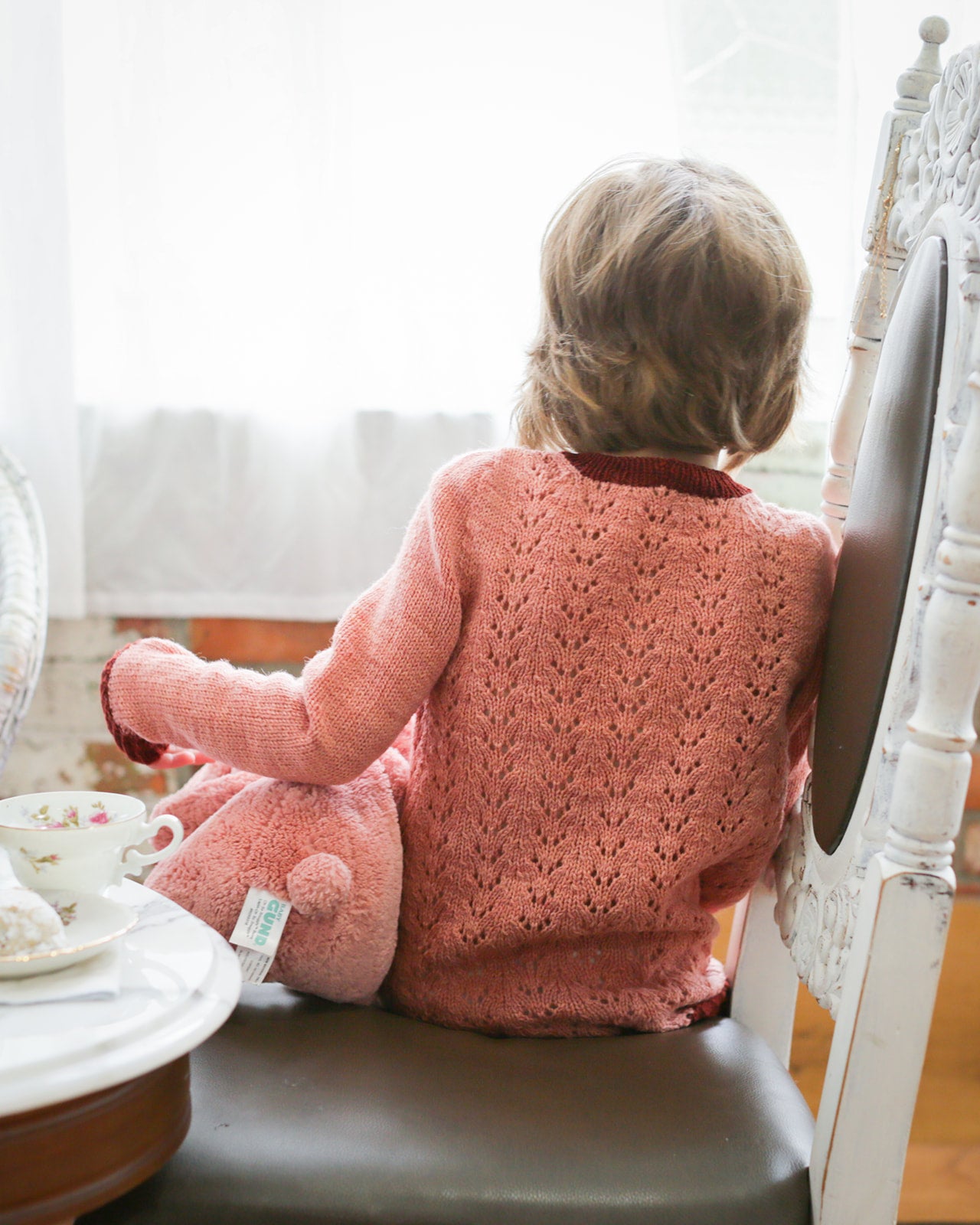 A young girl, seen from behind, sits on a white antique chair. She wears a light pink sweater, knit with a lace body and a dark pink collar, cuffs and hem. Teacups and a pink stuffed animal can be seen to her side.