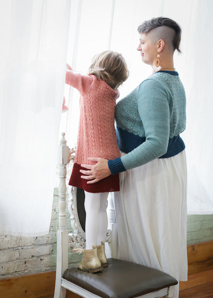 A young girl stands on a white antique chair, wearing a dress length version of the Kid's Classic sweater. It's knit in light pink yarn with a lace design on the body, and a dark pink neckline and hem. It's been styled with white tights and gold boots. Beside her, Jen wears a white skirt and a cropped version of a matching sweater, knit in light blue with dark blue cuffs, neckline, and hem.