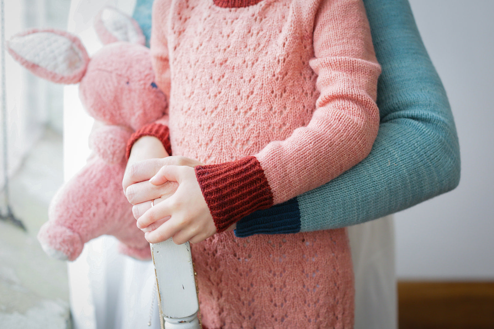 Seen from the shoulders down, a young girl, the arm of her mother around her, wears a long version of the Kid's Classic sweater. It's knit from pink yarn and dark pink cuffs. Her mother wears a matching light blue sweater with dark pink cuffs, and holds a pink stuffed animal.