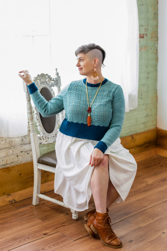 Jen sits on a white antique chair, turned partially away from the camera. She wears a white skirt and brown boots with a lace knit sweater. The sweater is made with light blue yarn and a dark blue hem, cuffs, and neckline.