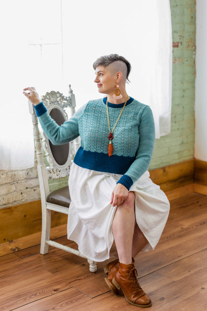 Jen sits on a white antique chair, turned partially away from the camera. She wears a white skirt and brown boots with a lace knit sweater. The sweater is made with light blue yarn and a dark blue hem, cuffs, and neckline.