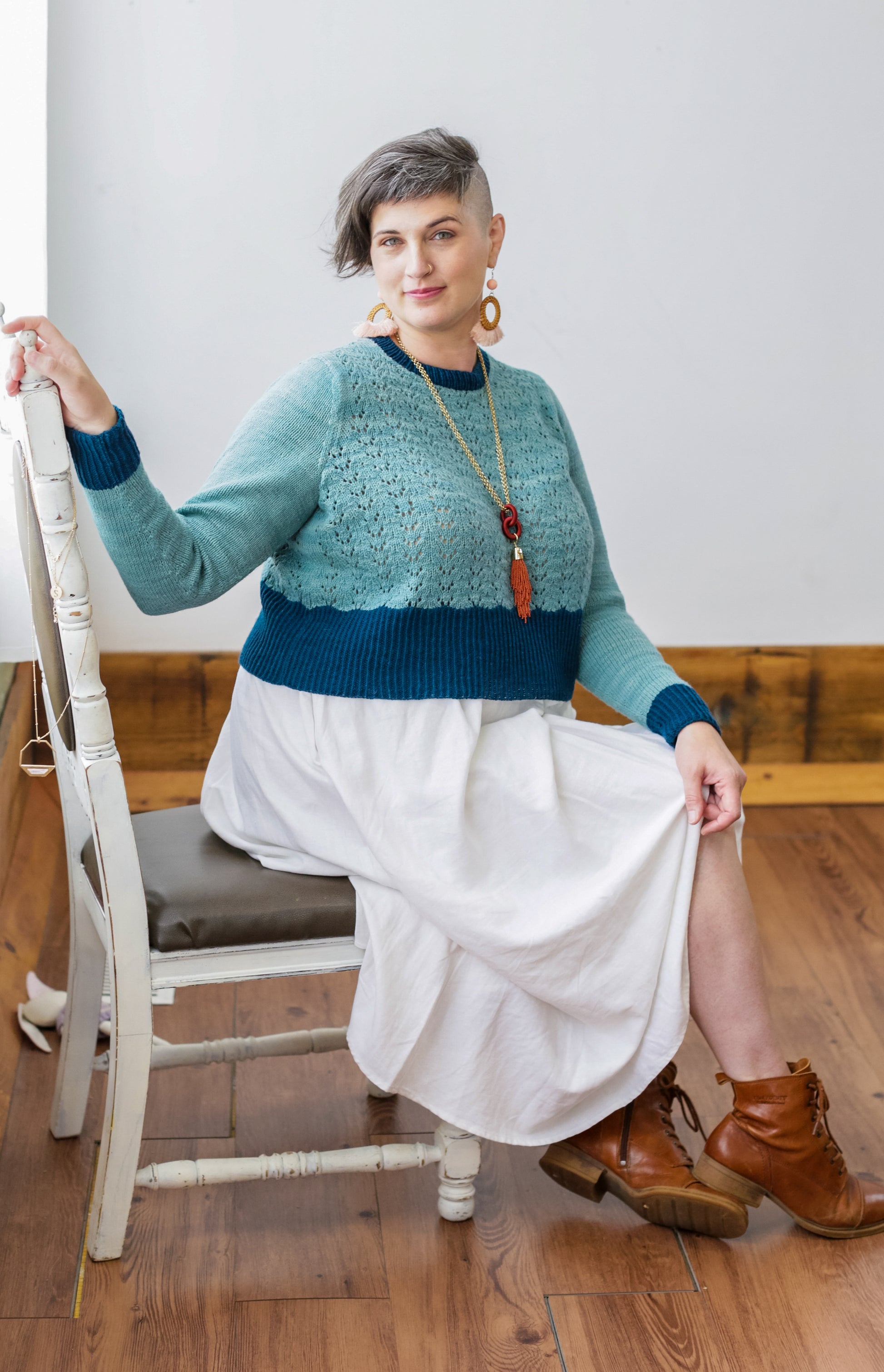 Seen from the side, Jen sits on a white antique chair. She wears a white skirt and brown boots with a cropped, lace knit pullover. The pullover is made with light blue yarn and contrasting dark blue cuffs, hem, and neckline.