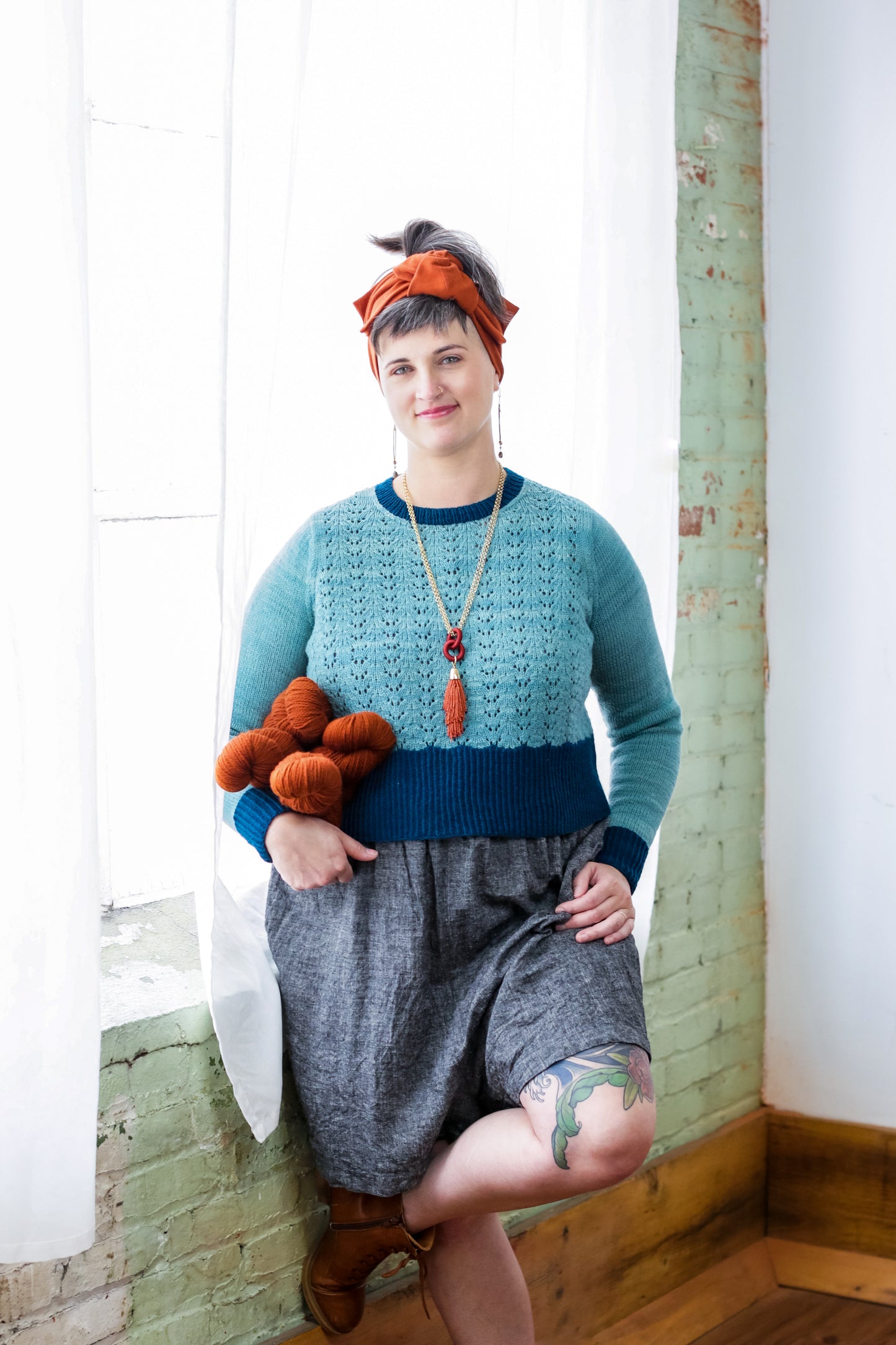Jen leans against a window ledge, holding four skeins of yarn and smiling at the camera. She wears a light and dark blue lace knit sweater, knit from her Classic Colorblock pattern. She's styled it with a grey skirt and brown boots.