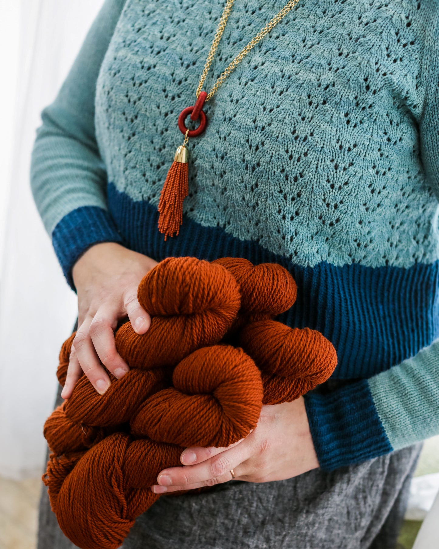 Seen close up, Jen wears a cropped pullover, knit from light blue yarn and featuring a lace design with dark blue hem, cuffs, and neckline. She holds four brick orange skeins of yarn that match her tasseled necklace.