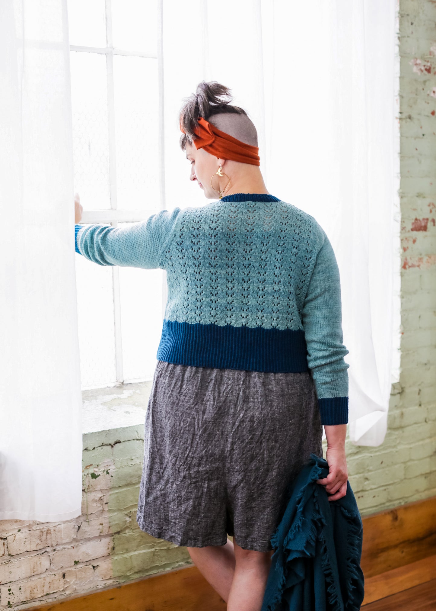Seen from behind, Jen wears a grey skirt with a light blue cropped pullover, featuring a lace design on the body and dark blue cuffs, hem, and neckline. She holds a matching dark blue shawl and stands in front of a window.