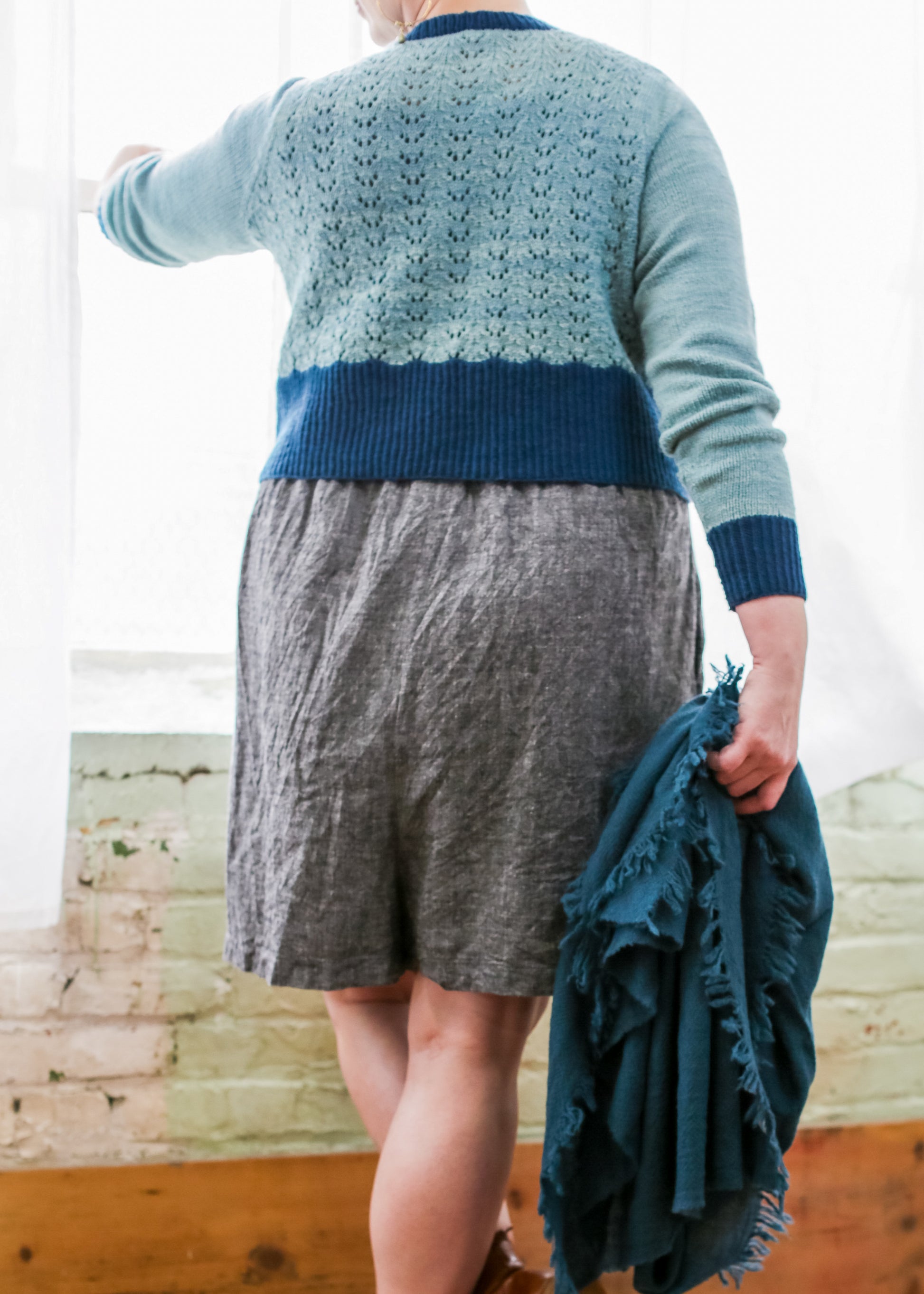 Seen from the shoulders down, Jen wears a grey skirt with a light blue cropped pullover, featuring a lace design on the body and dark blue cuffs, hem, and neckline. She holds a matching dark blue shawl and stands in front of a window.