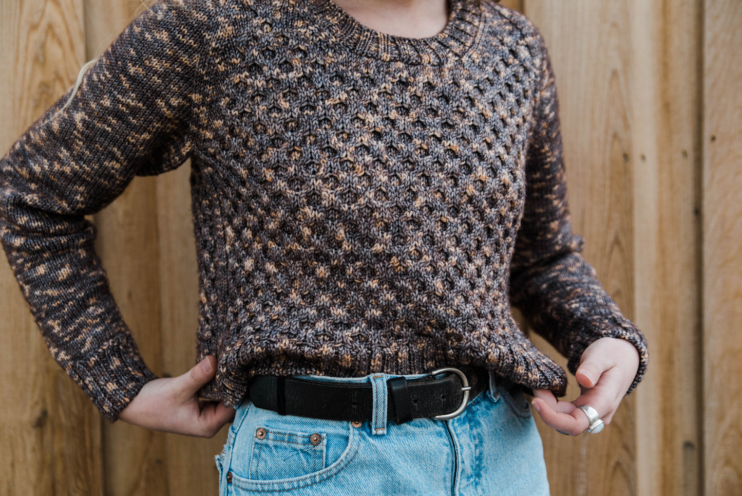 Haley, seen from the shoulder downs, tucks the edge of her honeycomb cable knit sweater into her blue jeans. The sweater is knit with brown, variegated yarn.
