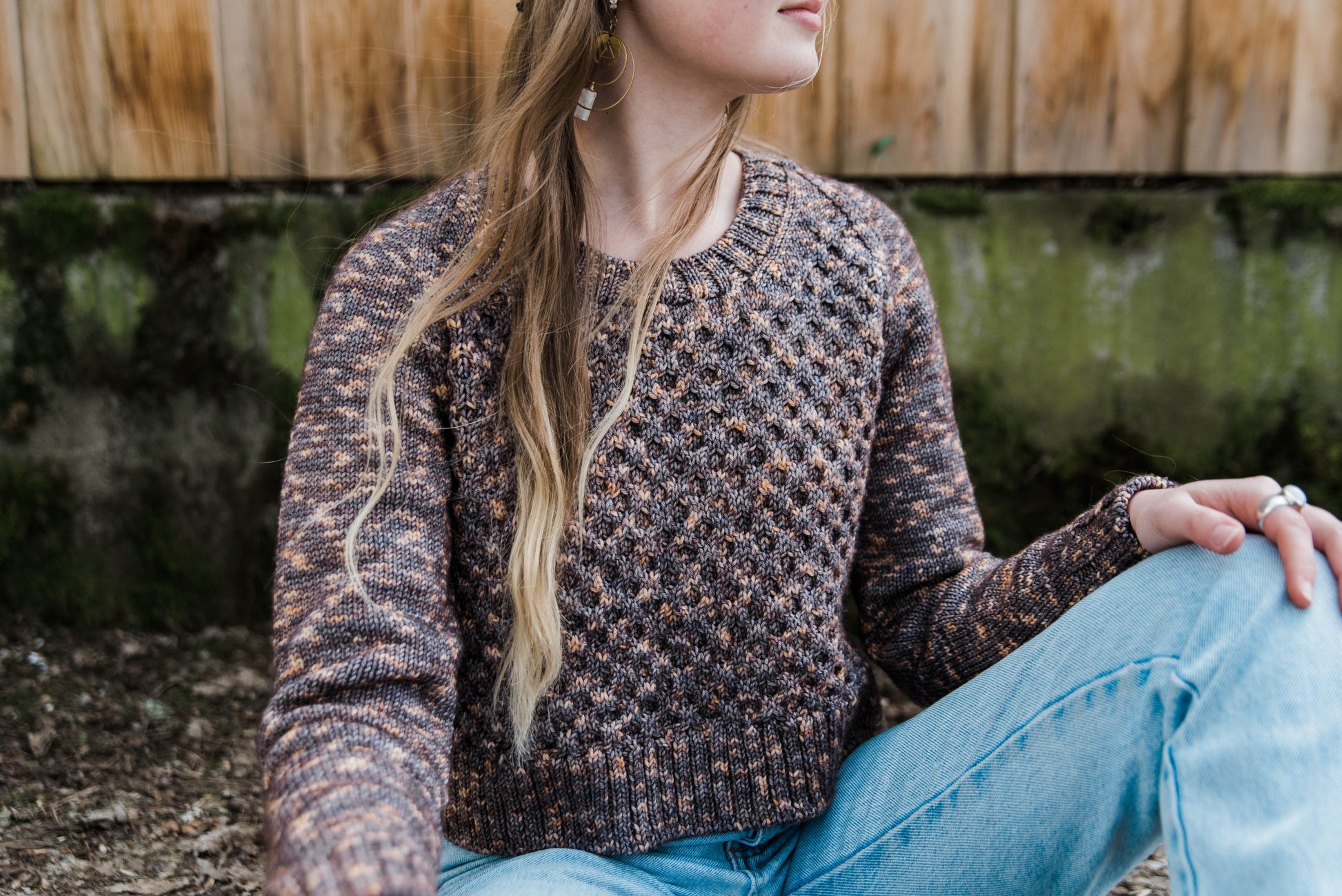 Haley, seen from the neck down, sits in front of a wooden fence. She wears blue jeans with a cropped knit sweater that features a honeycomb stitch pattern, knit in variegated brown yarn.