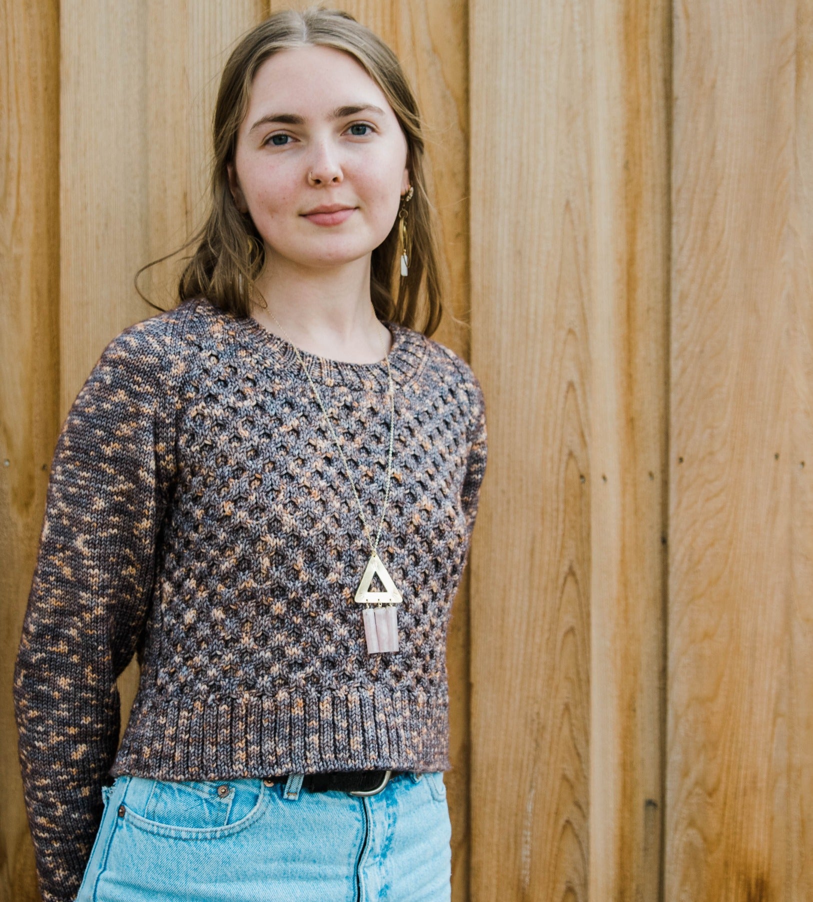 Haley smiles at the camera, wearing blue jeans with a cropped knit pullover. The pullover, Herbalist, features a honeycomb stitch pattern on the body and is knit in variegated brown yarn.